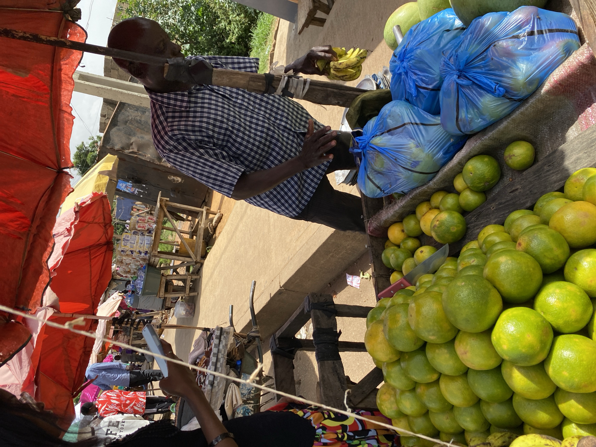 A man, likely a vendor, stands in a marketplace in Bukuru, Nigeria. He is wearing a blue and white checkered shirt and dark pants. He holds a bunch of bananas in his left hand. In front of him, on a wooden stall, are several large blue plastic bags filled with oranges. More oranges are displayed loose on the stall. A knife with a red handle is visible among the oranges. In the background, there are market stalls with red awnings and shelves stocked with various goods, including bottled water. The scene appears to be outdoors, under daylight, with some trees visible in the upper right corner. The ground appears to be made of concrete or packed earth.
