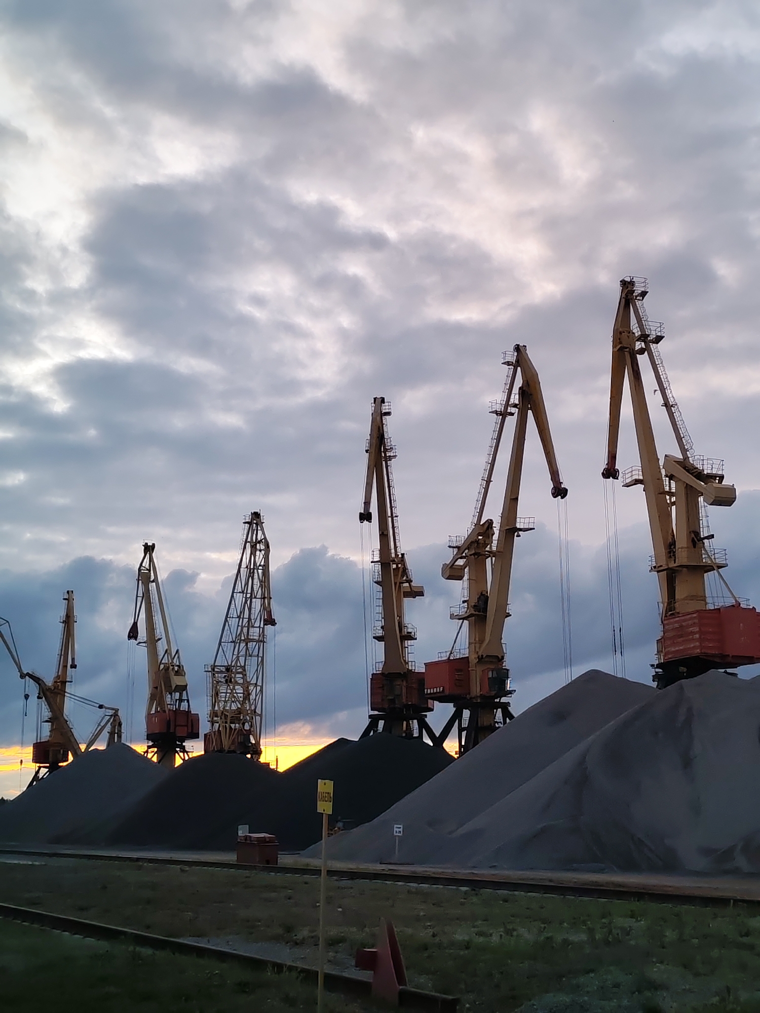 The image depicts a view of a port in Rybinsk, Russia, under a cloudy sky with hints of sunset peeking through. In the foreground, there are piles of dark gray gravel or sand, with railway tracks and a grassy embankment running horizontally across the bottom of the frame. Several large, yellow gantry cranes are positioned behind the piles of material. The cranes vary in size and design, with some appearing more industrial and skeletal than others. One crane on the far right has a red hopper attached. In the mid-ground, a yellow sign with Cyrillic lettering is visible, partially obscured by a pile of gravel. The overall scene conveys a sense of industrial activity and the vastness of the port operations. There are no people visible in the image.