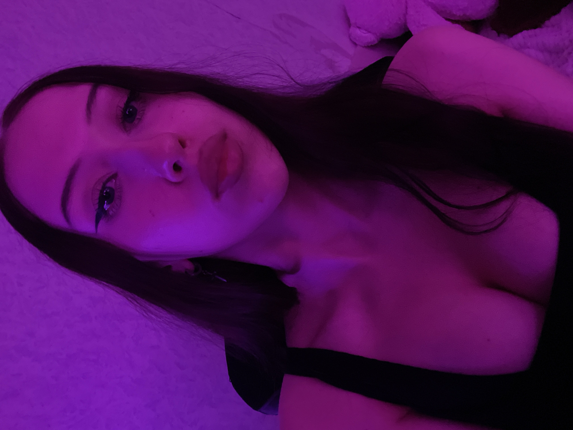 The image shows a young woman with long dark hair and winged eyeliner. She is wearing a black top that reveals her décolletage. The scene is illuminated with purple lighting, casting a strong glow on her skin and creating a vibrant, almost surreal atmosphere. The background appears to be a textured wall, also lit in purple. There are hints of what might be a stuffed animal on the right side of the frame, also bathed in the purple light. No specific location in Votkinsk, Russia, can be determined from the image itself.