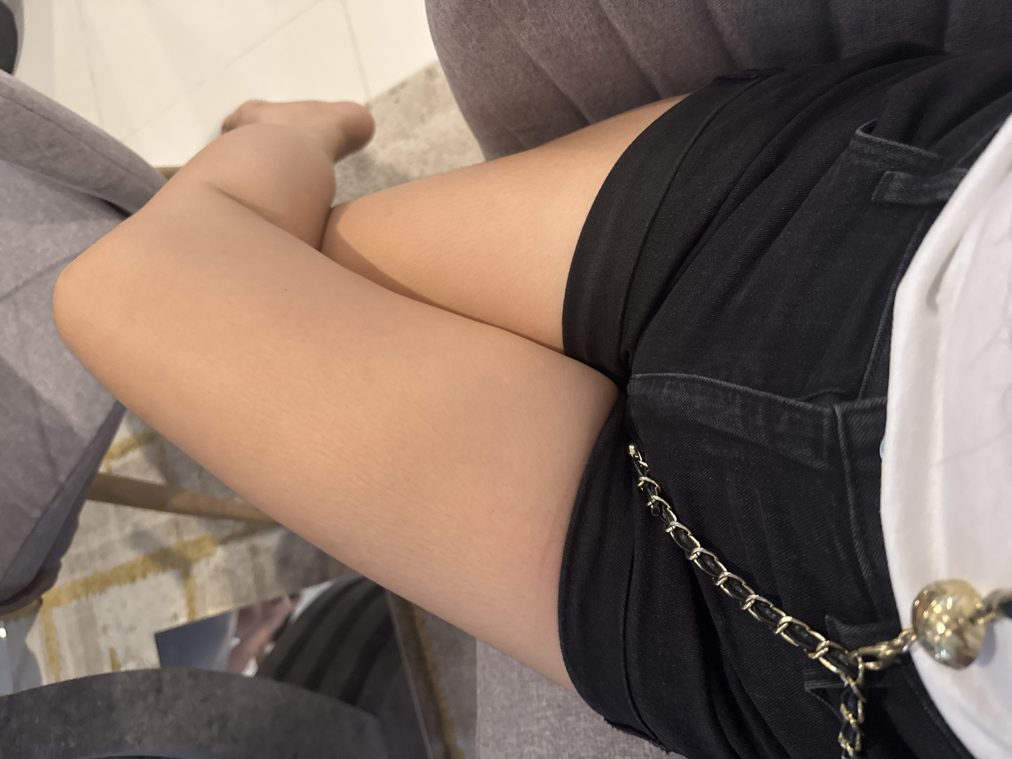 The image shows a close-up view of a person's legs crossed, wearing black denim shorts and a white top. A gold chain with a circular charm hangs from the shorts. The person is seated on a gray upholstered chair, with a patterned rug visible on the floor behind them. The lighting suggests an indoor setting. There is no identifiable information within the image that can place the location specifically in Davao City, Philippines.