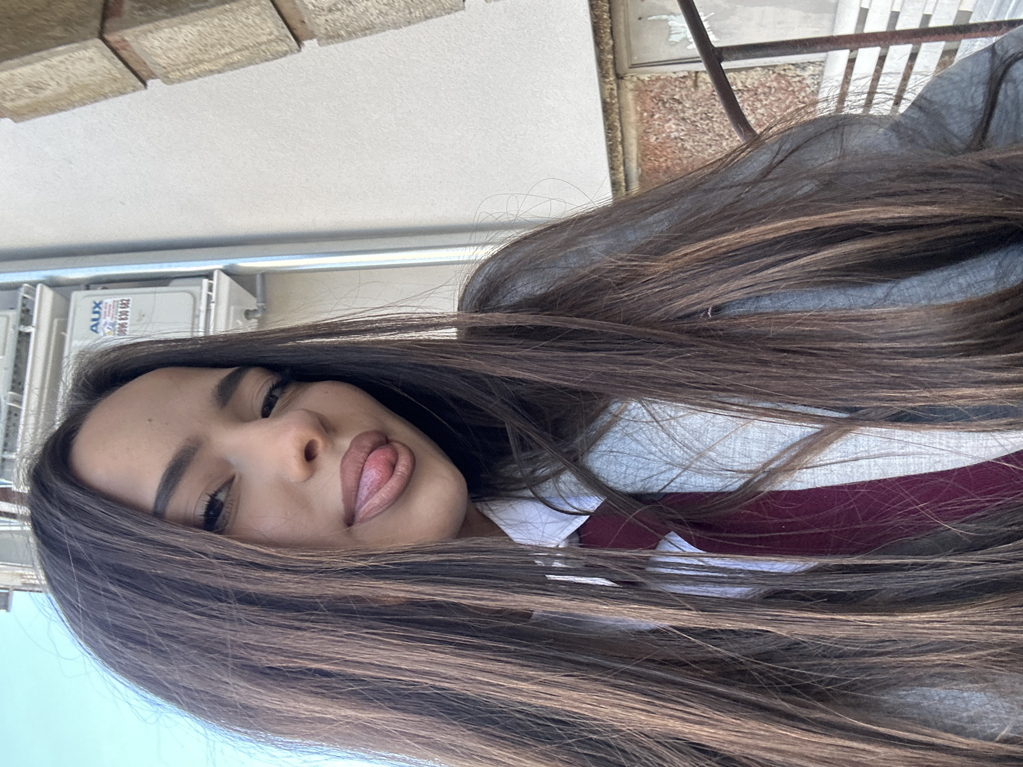 A medium shot depicts a woman with long, dark brown hair, facing the camera with her head tilted to the left. She is wearing a white collared shirt with a maroon tie underneath a gray hoodie. Her lips are slightly parted, revealing a portion of her tongue. Behind her, a beige brick wall is visible at the top of the frame, and an exterior wall with air conditioning units is to the left. A metal railing can be seen in the upper right corner of the image. The location is in an unknown city.
