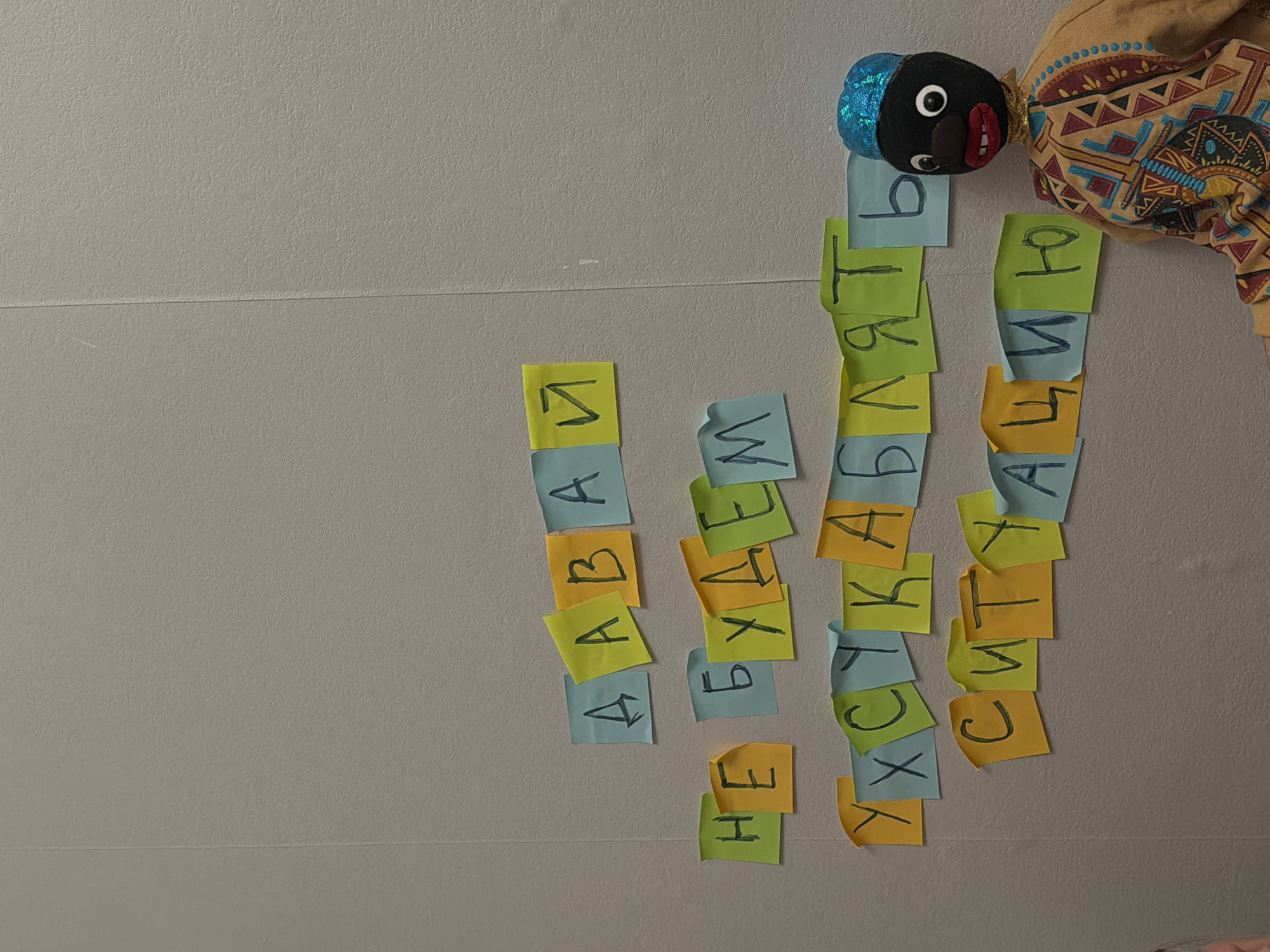 A handmade doll with a dark brown face, a sequined blue cap, and a colorful robe is affixed to a gray wall. Vertical rows of sticky notes displaying Cyrillic letters are aligned adjacent to the doll. The letters appear to spell out words in Russian. The overall scene suggests a learning activity involving the Cyrillic alphabet and potentially related to Perm, Russia.