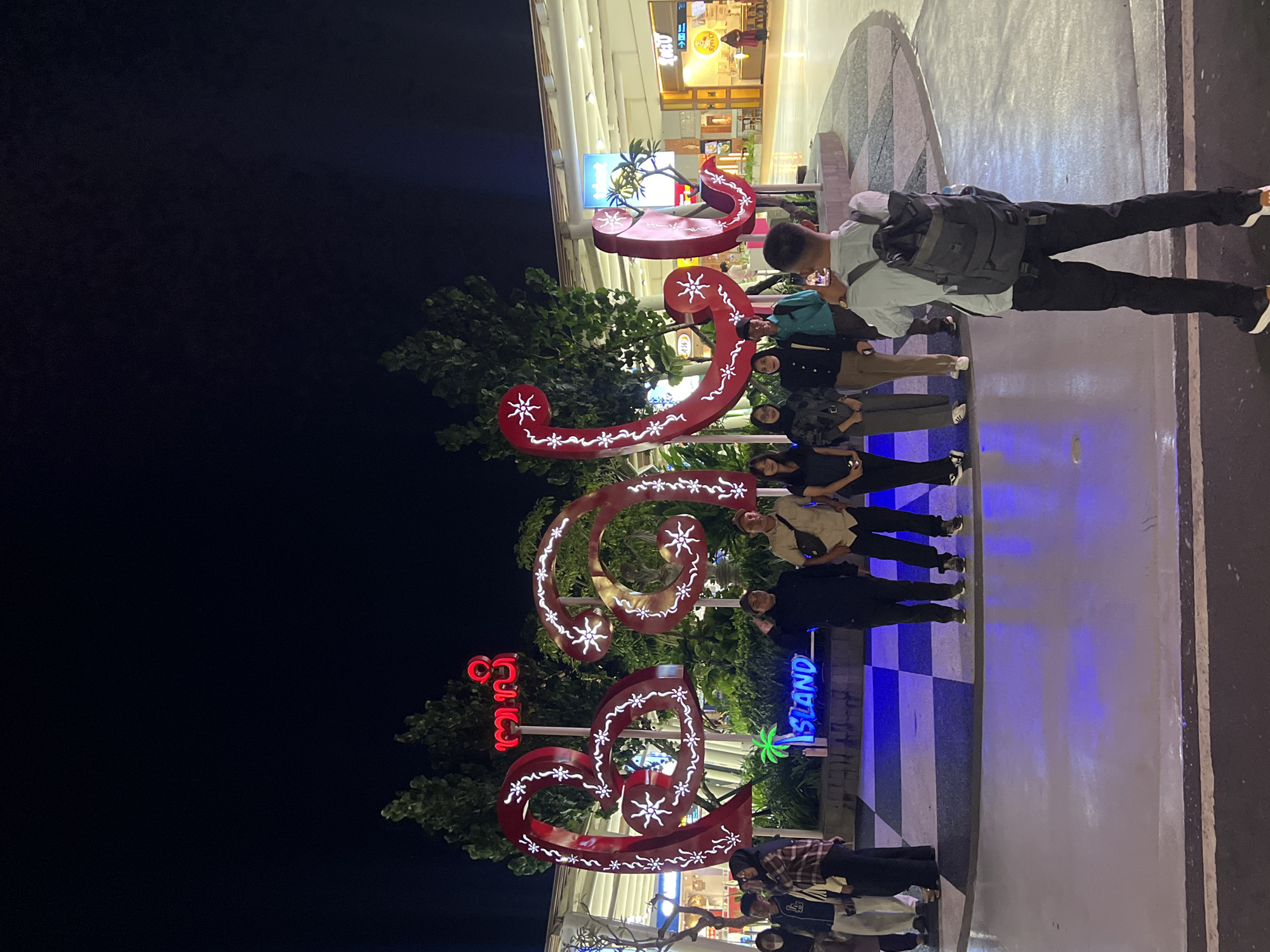 The image captures a group of people posing at night in an illuminated outdoor setting in Kuta, Indonesia. The scene appears to be a public area or the entrance to a commercial complex, brightly lit against a dark night sky.
In the foreground, approximately seven to eight individuals are visible, most of whom are looking towards the camera, suggesting they are posing for a group photograph. One person in the center-right of the frame, wearing a light-colored shirt and a dark backpack, has their back to the camera. The individuals are dressed in casual attire, indicating a relaxed social outing.
The most prominent elements are three large, red, ornate decorative structures. These structures are highly stylized, resembling abstract letters or symbols, possibly in a local script. They are adorned with numerous glowing white snowflake-like patterns and undulating white lines that stand out vibrantly against the red. Behind and around these decorations, lush green trees and foliage are visible, adding to the outdoor ambiance.
To the right of the decorative structures, a distinct blue neon sign spells out "ISLAND" and features a stylized palm tree icon, clearly indicating a tropical location. The ground surface is a mix of a black and white checkerboard pattern and a reflective paved area, illuminated by various light sources. In the background, brightly lit modern buildings with multiple storefronts can be seen, though specific shop names are largely indistinct, with one sign possibly reading "O'Bao." The overall atmosphere is festive and lively, consistent with an evening leisure activity in a popular destination.