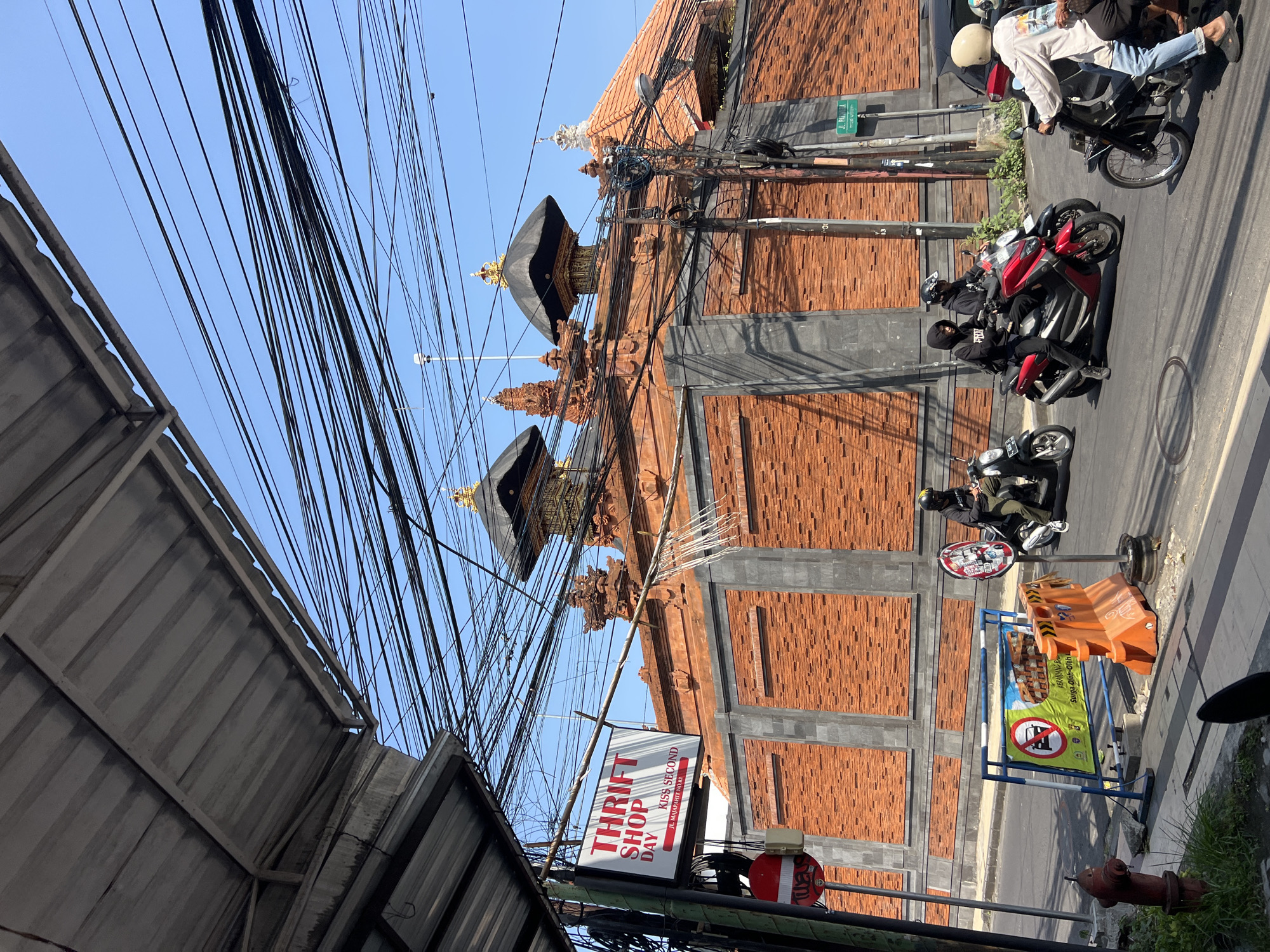 In Kuta, Indonesia, a sun-drenched street scene unfolds under a canopy of tangled electrical wires. A brick wall with gray stone accents and decorative spires forms the backdrop, leading to buildings with traditional tiled roofs. In the foreground, a group of motorcyclists navigates the road. On the left, a sign for "THRIFT SHOP DAY 1 KISS SECOND" is mounted on a pole next to a red and white traffic barrier. Further down the street, orange construction barriers and a blue-framed sign with an image of goods and text in Indonesian are visible. The road is paved, and a red fire hydrant stands on the sidewalk. The sky is clear and blue.