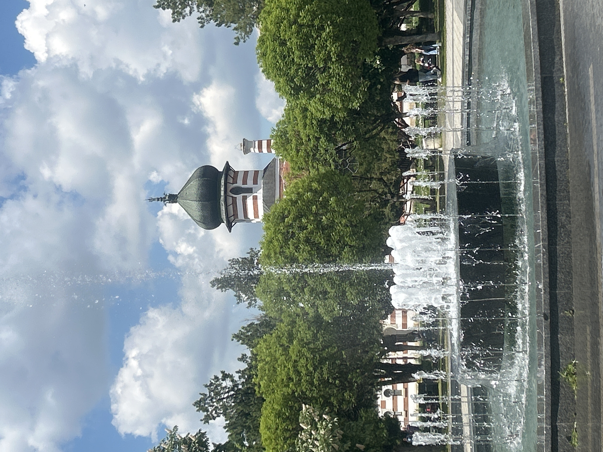 The image depicts a fountain in Pleven, Bulgaria, with a historic building in the background. The fountain is actively spraying water, creating a visual barrier and obscuring the view. The building features a dome and red-and-white striped sections. Trees and foliage surround the area. A group of people is visible in the background, near the fountain. The sky is cloudy.