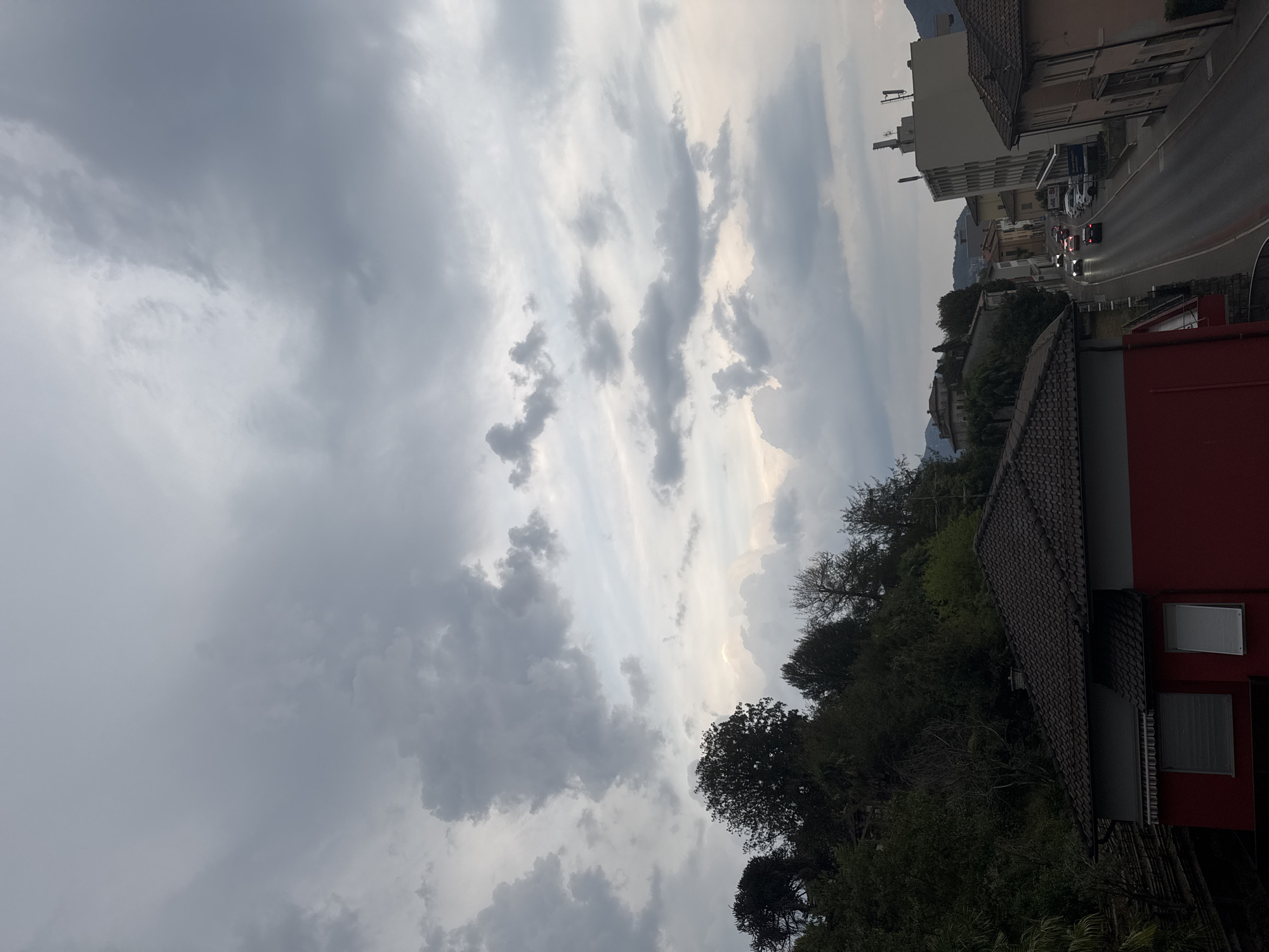 This is an outdoor, eye-level, medium shot of a street in Balerna, Switzerland, under a cloudy sky. The image captures a residential area with a street running through it. On the left, a red building with a dark tiled roof is partially visible in the foreground. To the right of this building, the street curves, with several cars driving on it. Buildings line both sides of the street, varying in height and architectural style. Trees and vegetation are present on a hillside behind the buildings on the left. The sky is predominantly filled with grey clouds, with some lighter patches suggesting breaks in the overcast. No people are visible in the scene. The overall impression is that of a typical European town street on a cloudy day.