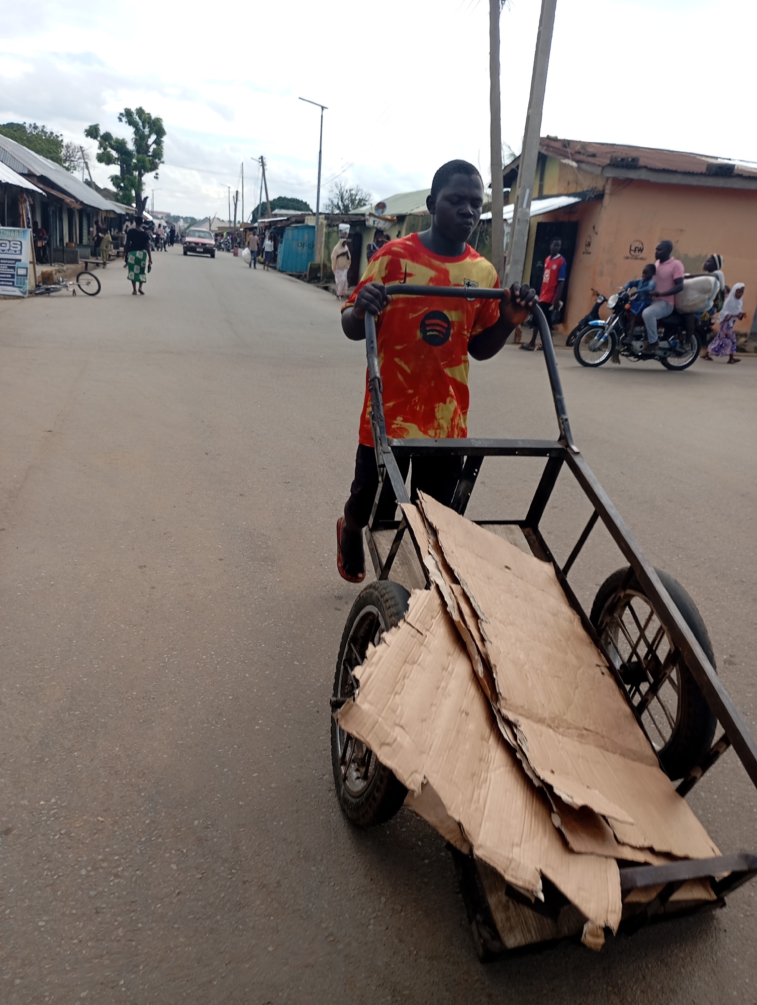 A young Black male is pushing a metal handcart laden with flattened cardboard boxes down a paved street in Shendam, Nigeria. He is wearing an orange t-shirt with a yellow and green design, black pants, and dark shoes. The handcart has two large wheels and a frame that extends upwards. In the background, other people are visible, including a man riding a motorcycle with two passengers, and individuals walking along the sides of the street. Buildings with corrugated metal roofs line the street, and trees and utility poles are also present. The sky is overcast. The scene appears to be a commercial or residential area within Shendam.