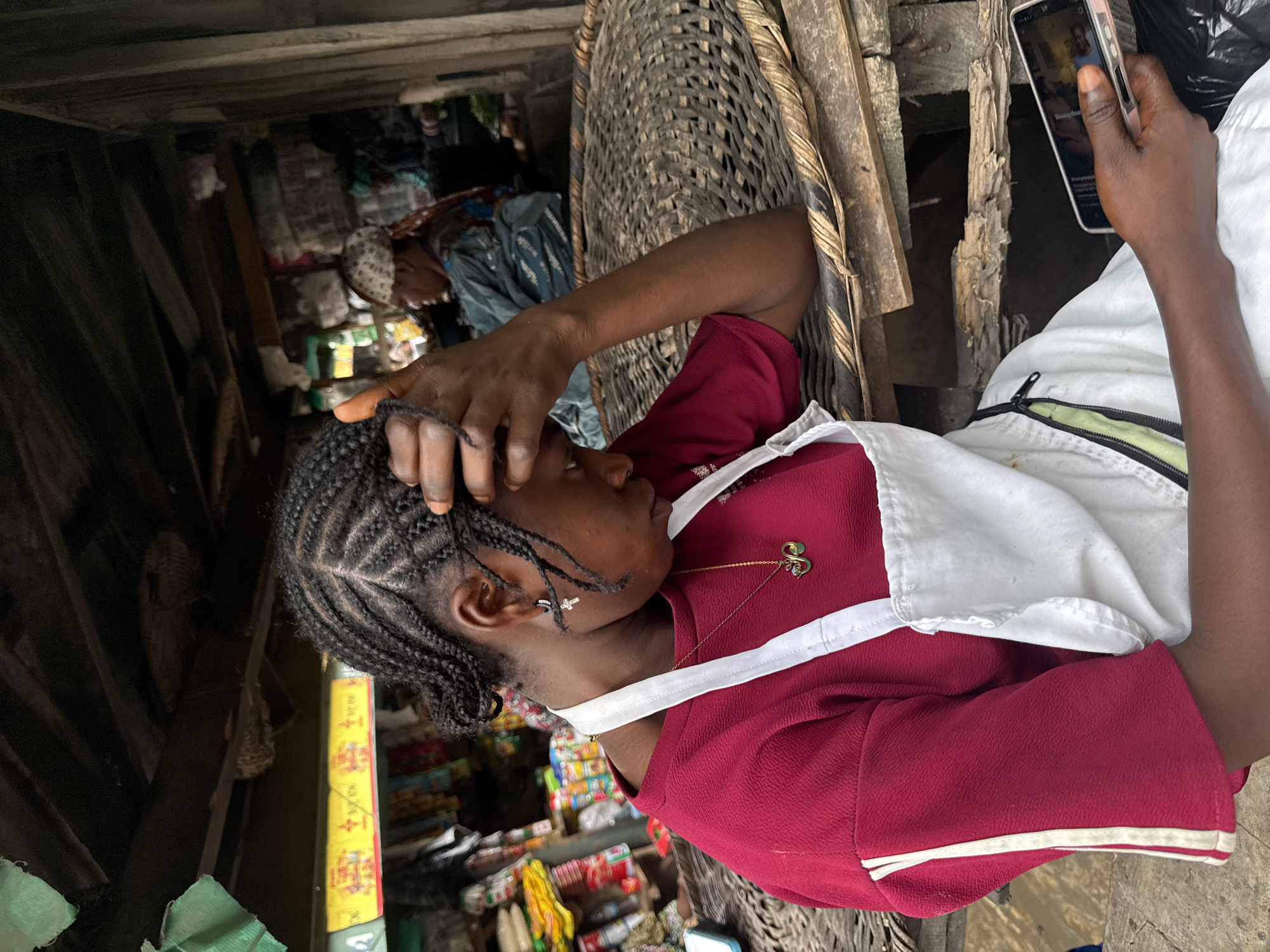 In a market stall in Lagos, Nigeria, a young woman with braided hair rests her forehead on her hand, looking down at a smartphone she holds. She wears a red shirt with white stripes on the sleeves and a white apron. A gold necklace with a green charm is visible on her neck. Another woman in the background, wearing a patterned headscarf, is seated behind a counter with goods. The stall is constructed with rough wood and thatched roofing, with shelves stocked with various items, including packaged goods and fabrics, lining the walls. A large woven basket sits on a wooden surface in the foreground. The scene appears to be a typical day in a bustling Nigerian marketplace.