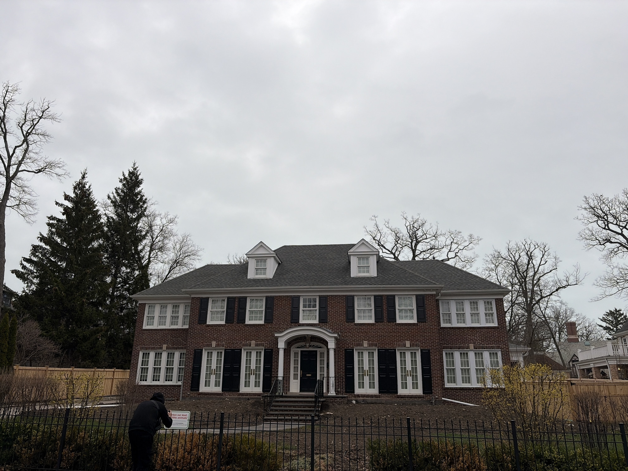 The image depicts the exterior of a large, two-story brick house located in Winnetka, Illinois, USA. The house has a dark grey roof with three dormer windows. The front facade features symmetrically arranged windows with black shutters and white trim, flanking a central doorway. A small porch covers the entrance.
A black iron fence runs along the front of the property, separating it from the street. Bare trees surround the house, suggesting a late fall or winter season. A person stands near the fence, possibly taking a picture. A sign is attached to the fence. The sky is overcast. A wooden fence can be seen in the background to the right of the image.