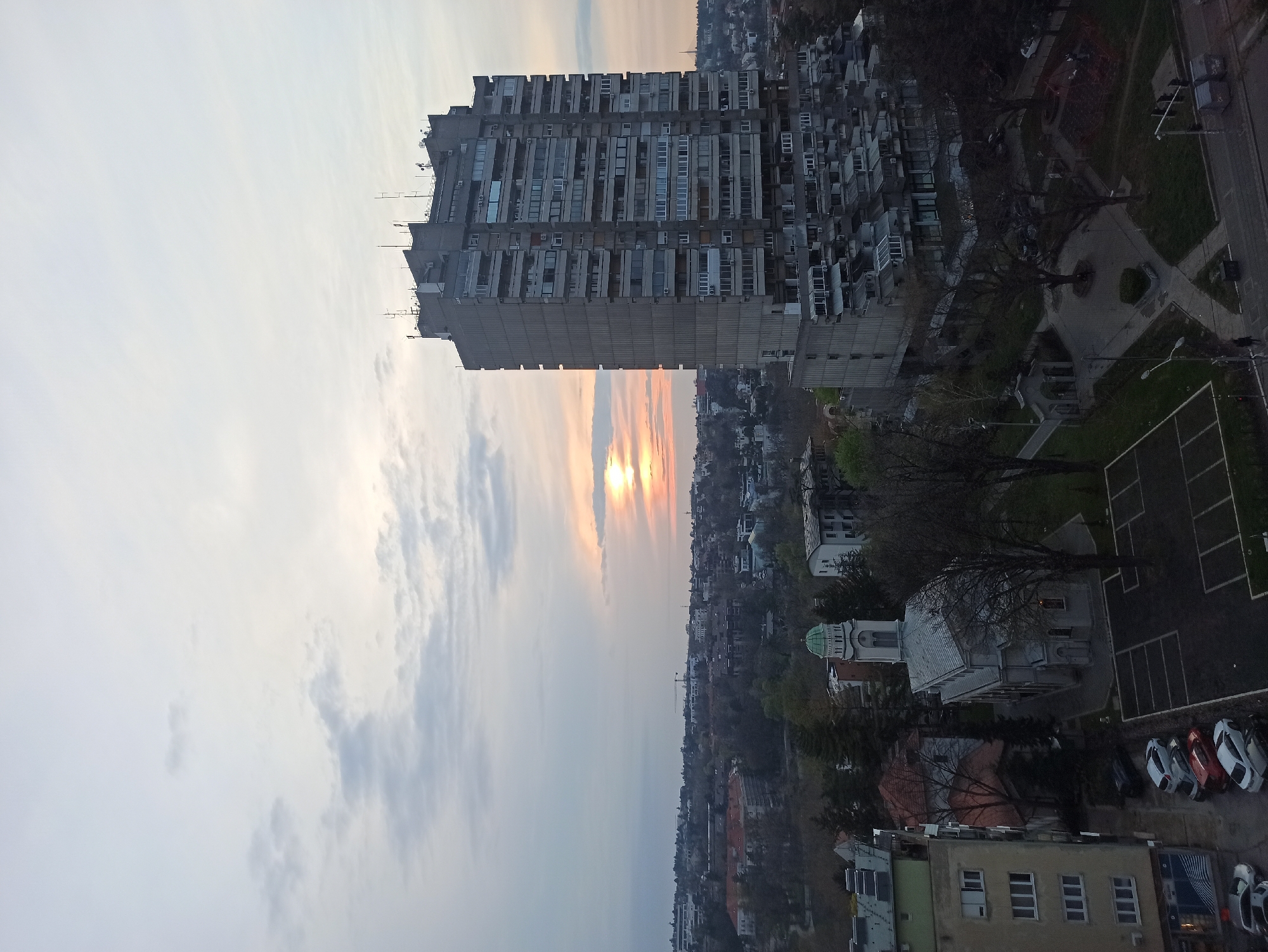 The image presents an elevated view of a cityscape at sunset, dominated by a prominent, tall, concrete apartment building in the foreground. This building, with its repetitive grid of balconies, is situated in the urban municipality of Vozdovac in Serbia. The sky transitions from a bright, setting sun to a cloud-streaked, dusky sky. Below the apartment building, the landscape is densely populated with buildings of various heights and styles. At the very bottom right, a building with a green-domed roof can be seen, along with a parking lot containing several cars. The general environment suggests a densely populated urban setting undergoing the transition from day to night. No people are visible in the image.