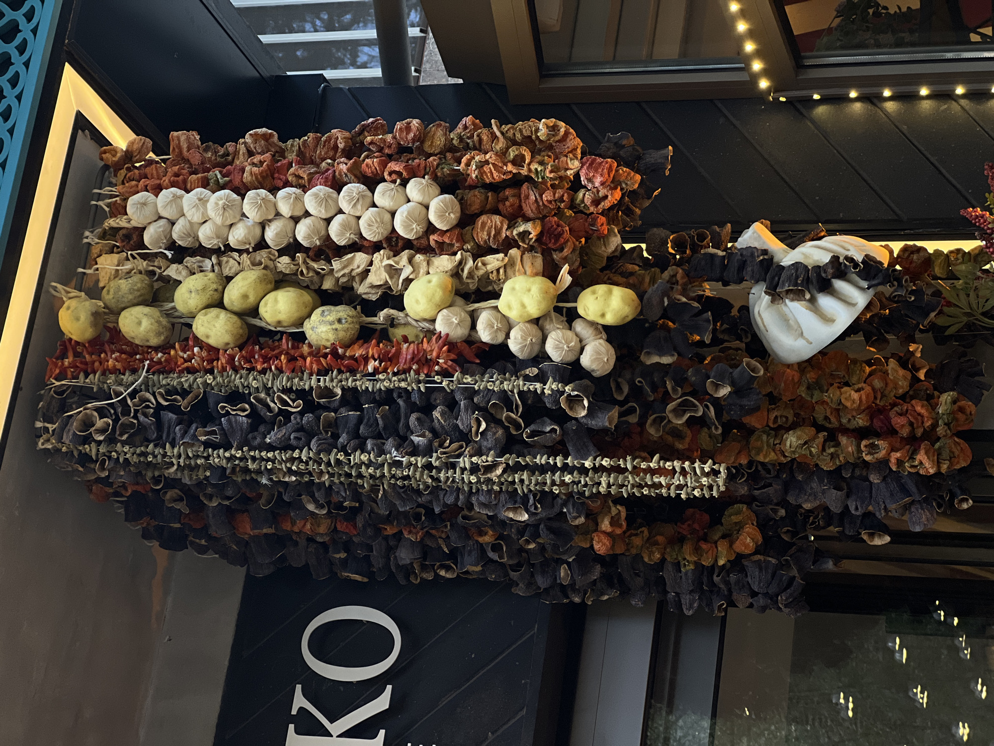 The image displays a vibrant arrangement of various dried vegetables and fruits, traditionally strung together, likely as a decorative display outside a shop or restaurant in Yerevan, Armenia.
The main subjects are numerous strings of produce hanging vertically and horizontally. Prominently featured are strings of dried red peppers or tomatoes, characterized by their reddish-brown, shriveled appearance. Below these, there are strands of dried garlic bulbs, appearing as white, round clusters. Further down, strings of what look like dried potatoes or small gourds, yellow-green in color, are visible. Darker, purplish-black dried eggplants are also extensively featured, strung up in multiple rows. Additionally, finer strings of small, dried chili peppers can be seen. To the right, a large, lighter-colored gourd or decorative item is adorned with dried eggplant shells.
The items are arranged in neat, horizontal rows, creating a textured and colorful facade. The setting appears to be outdoors, beneath an overhang or awning of a building, with a window reflecting ambient lights visible in the background, suggesting it is evening or night. String lights are visible along the top edge of the structure, further indicating artificial illumination.
A partial sign with the letters "KO" in white font against a dark background is visible at the bottom left of the image. The overall impression is one of traditional abundance and rustic charm, common in Armenian and regional culinary traditions for preparing winter provisions or for decoration.