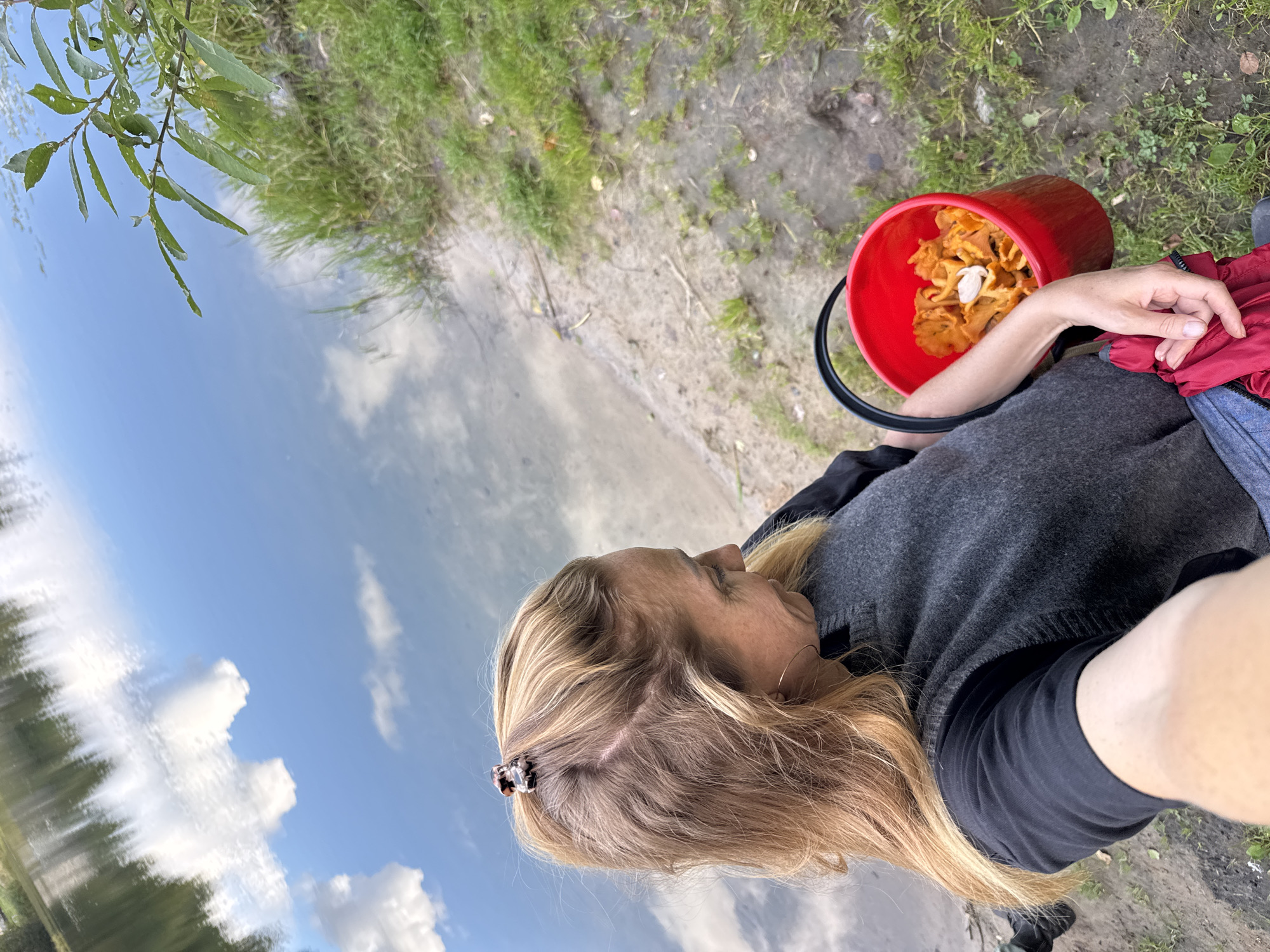A woman with blonde hair pulled back with a clip is holding a red bucket full of chanterelle mushrooms. She is wearing a dark long-sleeved shirt and a gray vest. The woman is standing on the sandy bank of a body of water, possibly a lake or river, with green grass and a tree branch visible in the background. The water reflects the blue sky and white clouds. The scene appears to be outdoors in a natural setting, possibly a park or forest, located in the city of Mars, Russia.