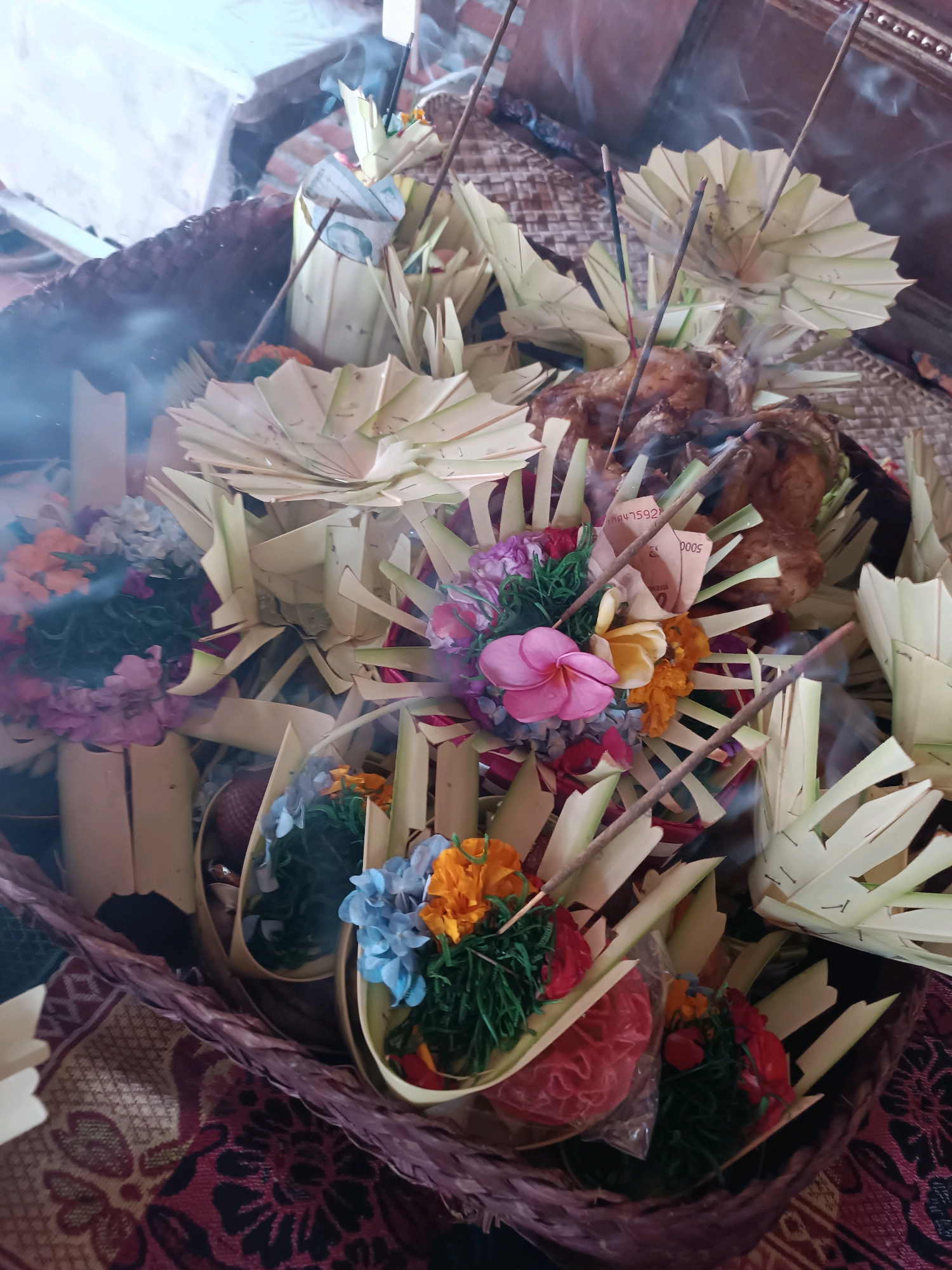 The image depicts a close-up view of several Balinese offerings, known as canang sari, arranged in woven baskets. The offerings are elaborate, featuring fresh flowers in various colors such as pink, yellow, orange, and blue, interspersed with green foliage. Many of the offerings are adorned with folded palm leaves, creating a fan-like structure. Incense sticks are visibly lit, with trails of smoke rising from them, indicating a ritual or ceremonial context. Some of the offerings also include pieces of currency, suggesting they are a form of prayer or thanksgiving. The background is slightly out of focus, but it appears to be an indoor or sheltered setting, with woven mats or textiles visible beneath the baskets. Based on the cultural elements present, the scene is likely located in Selemadeg, Bali, Indonesia, a region known for its traditional religious practices. There are no people visible in the image.