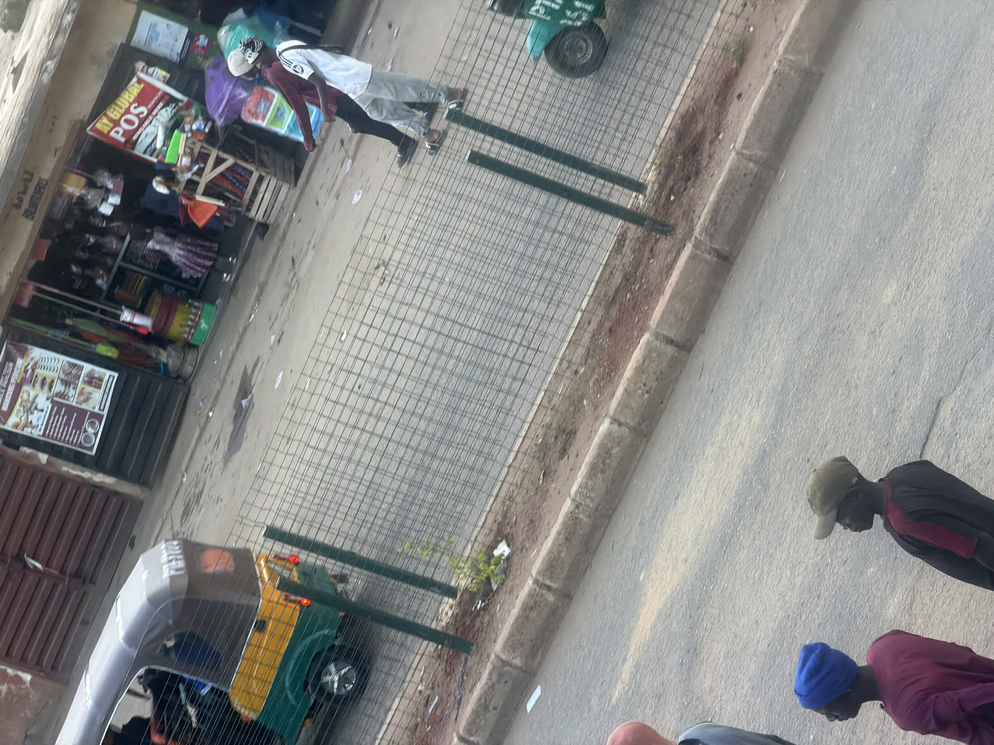 The image depicts a street scene in Bukuru, Nigeria, likely during the daytime with bright sunlight casting shadows. In the foreground, three individuals are visible from behind, one wearing a blue beanie and a maroon shirt, another in a green cap and a dark shirt with red accents, and a third partially obscured person. They appear to be standing on a paved road.
The midground shows a wire fence separating the road from a sidewalk or an area adjacent to shops. Behind the fence, a three-wheeled auto-rickshaw, commonly known as a "Keke Napep," is parked. The Keke is green and yellow. Further in the background, a small storefront is visible, with a sign that reads "IN GLOBAL POS" in red lettering on a yellow background. The shop appears to be selling various goods, including clothing and what looks like household items. Some people are visible inside or around the shop, one wearing a white shirt and grey pants and another in a maroon long-sleeved shirt. Another person is wearing a headscarf. There is also a display of umbrellas and other items outside the shop.
The overall scene suggests a busy commercial street. The ground is paved, and there is some dirt and debris along the side of the road and near the fence. The lighting indicates it is daytime.