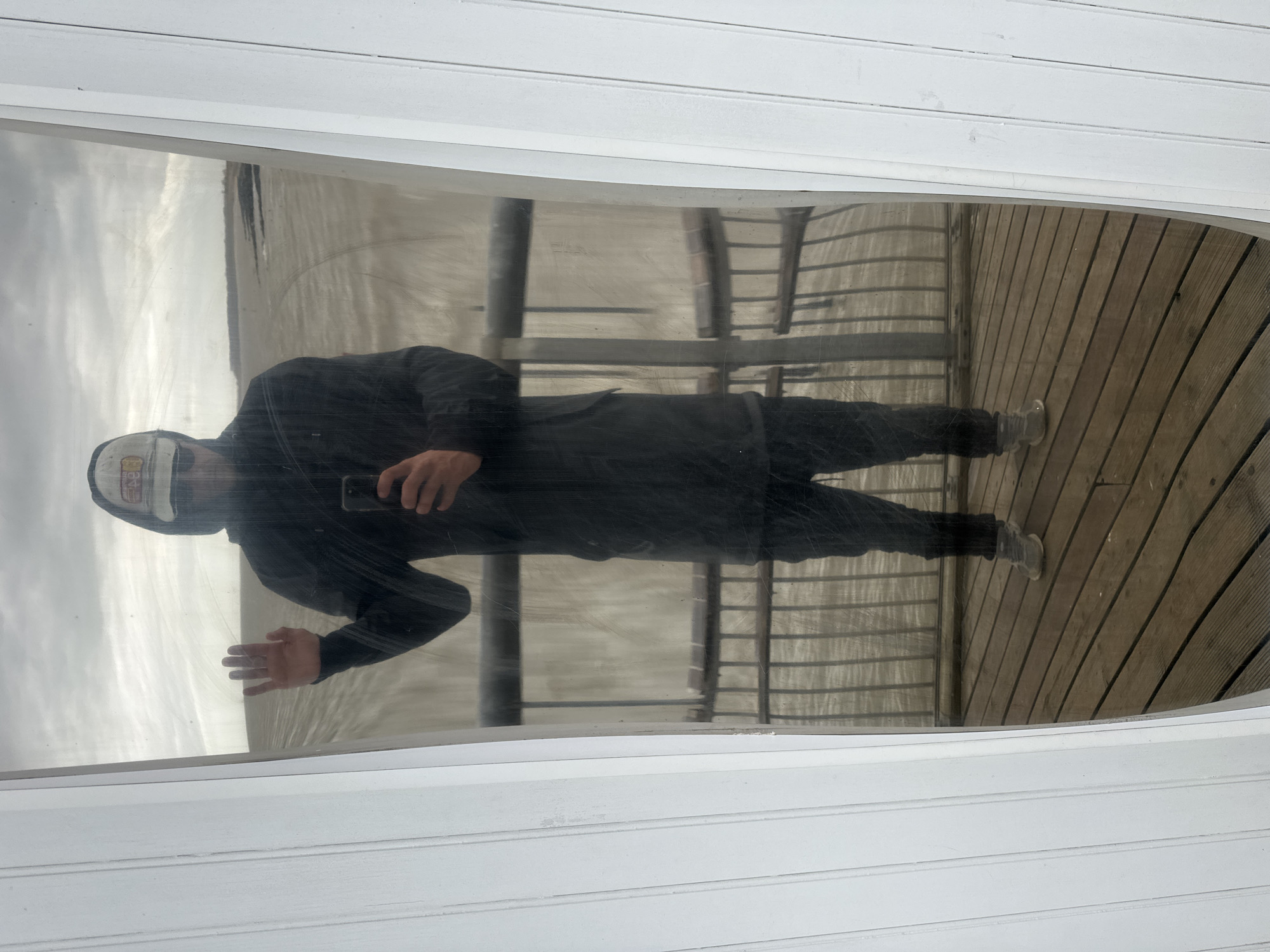 The image displays the reflection of a person in a large window or clear panel, offering a view of an outdoor environment. The person is dressed in dark clothing, comprising a hooded top and trousers, and wears a white baseball cap adorned with an unidentifiable, multi-colored logo on the front. They are holding a smartphone in their right hand, appearing to be taking the photograph of their own reflection. Their left hand is slightly raised, with the palm facing outwards.
The reflected background reveals a muted landscape. On the left side, a vast, grey expanse of either an overcast sky or a calm body of water is visible. On the right, a wooden deck or pier, constructed from horizontal planks, extends into the scene. Dark metal structural elements, possibly railings or supports, are also reflected. The entire reflected view is bordered at the top and bottom by white, horizontally paneled surfaces, suggesting the architecture of the surrounding building. The weather appears overcast, and the setting indicates a daytime, waterfront location, consistent with East Suffolk, United Kingdom.