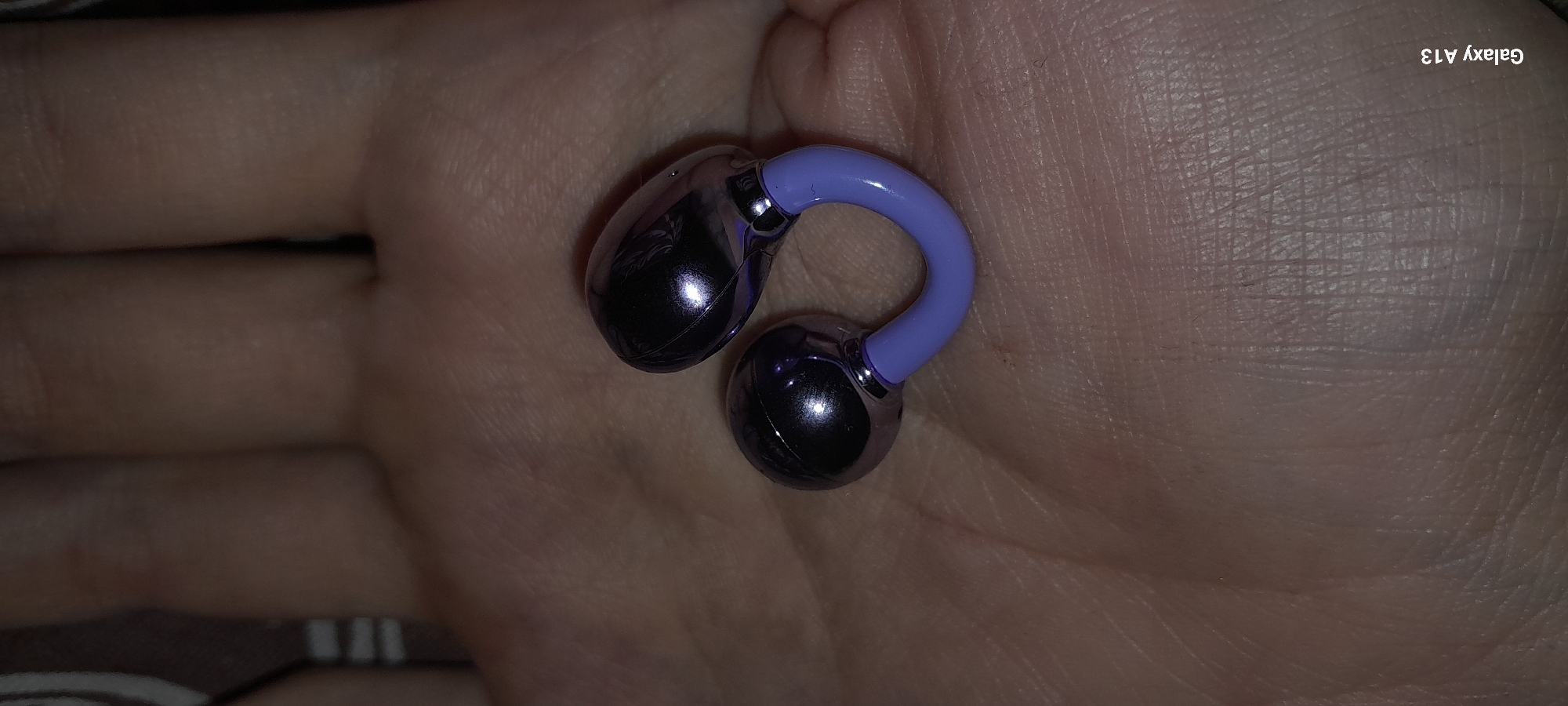 A close-up shot shows a purple and silver massage tool held in a person's hand. The tool has a u-shaped, lavender-colored middle section with two spherical, metallic silver ends. The hand appears to be resting on a surface with a subtle striped pattern. The background is blurred. The image is located in Novaya Razvodnaya, Russia.