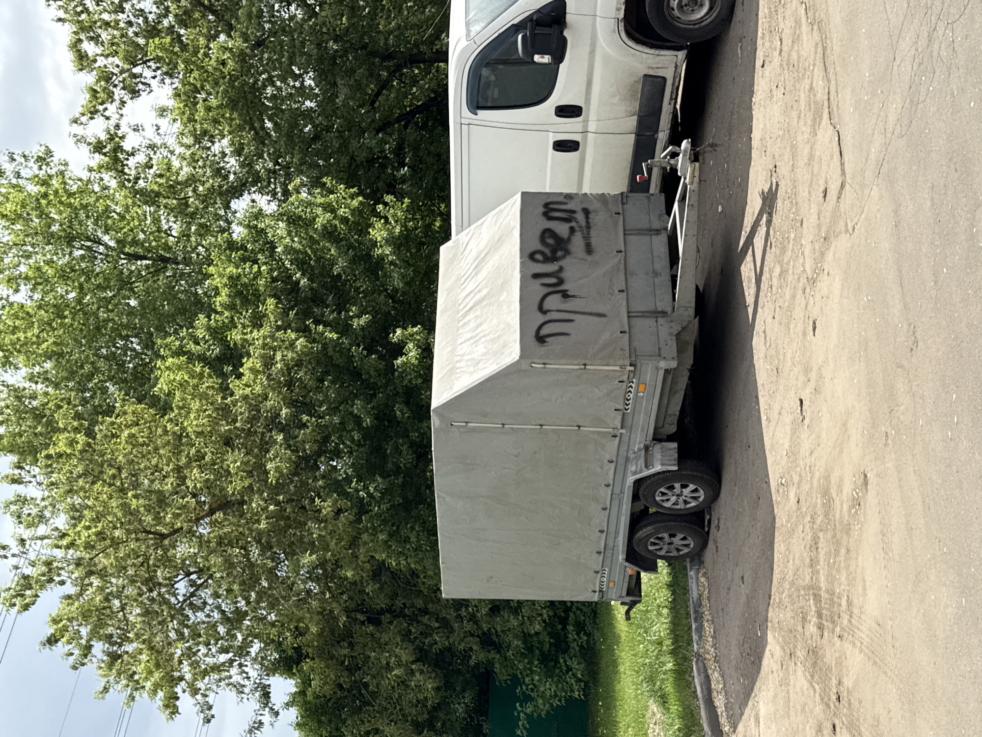 A white van and a light gray trailer are parked on a concrete sidewalk. The word "Привет" (hello) is written in black graffiti on the side of the trailer. A lush green tree is in the background. The image appears to be taken in Moscow, Russia, based on the Cyrillic writing and the overall urban environment.