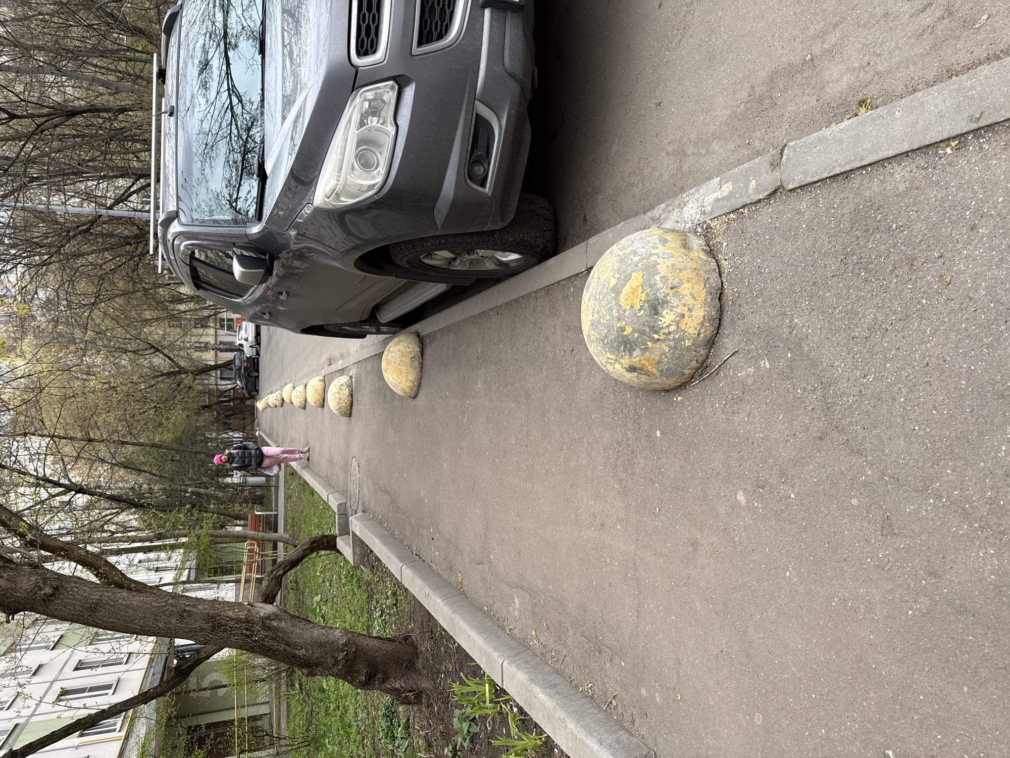 This image, taken in Moscow, Russia, captures a street scene from an elevated perspective, looking down a paved path alongside a road. In the foreground and extending along the left side of the frame, a dark gray SUV is parked, with its front end partially visible and its right front wheel resting on the curb. The vehicle has roof rails and side steps.
To the right of the parked car, a row of spherical, concrete bollards, some discolored with yellow and green patches, line the edge of a pedestrian path or sidewalk. The path is made of dark gray asphalt. Further down the path, a person, a woman, is walking away from the camera. She is dressed in a dark jacket, pink pants, and a bright pink beanie.
On the left side of the frame, beyond the car, are several bare deciduous trees, indicative of early spring foliage. A large tree with a thick, gnarled trunk is prominent in the immediate foreground on the left. Through the trees in the background, a multi-story, light-colored residential building (an apartment block) with numerous windows is visible. Two park benches, one appearing wooden and another with a yellow metal frame, are also seen in the distance near the building.
The lighting suggests daytime, possibly overcast, as there are no harsh shadows. The overall scene depicts a typical urban or suburban residential environment. No discernible text is visible on any objects in the image.