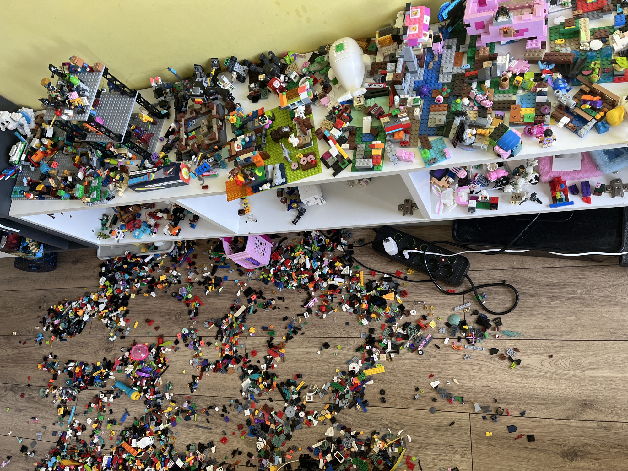 The image shows a messy room, possibly a child's playroom. A white shelf is crammed with assembled Lego creations and loose Lego bricks of various colors and sizes. A significant quantity of Lego bricks has spilled onto the wooden floor in front of the shelf, creating a scattered and chaotic appearance. A pink plastic basket sits amidst the spilled Lego bricks on the floor. A black power strip with several plugs and tangled wires is located near the shelf and the spilled Lego bricks. The background consists of a yellow wall. The location is inside a domestic building.