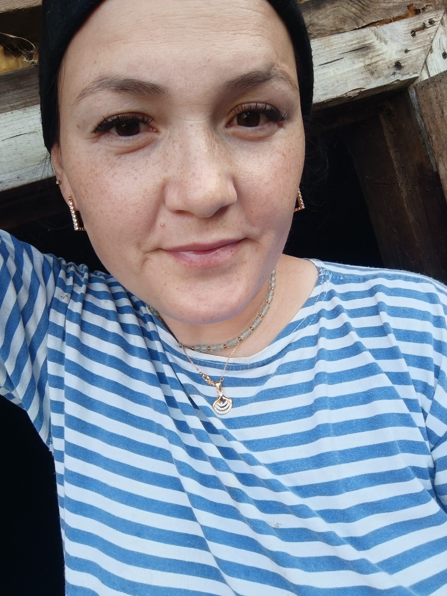 A selfie of a woman with freckles is taken from the chest up, with the woman looking directly at the camera. She is wearing a black beanie, a blue and white striped shirt, and a necklace with a shell pendant. She is also wearing square earrings with rhinestones. The background appears to be an outdoor setting with wooden structures and dark shadows. The image quality is good, with clear focus on the woman's face. There is no indication of the location being in the city of Karsha, Russia.