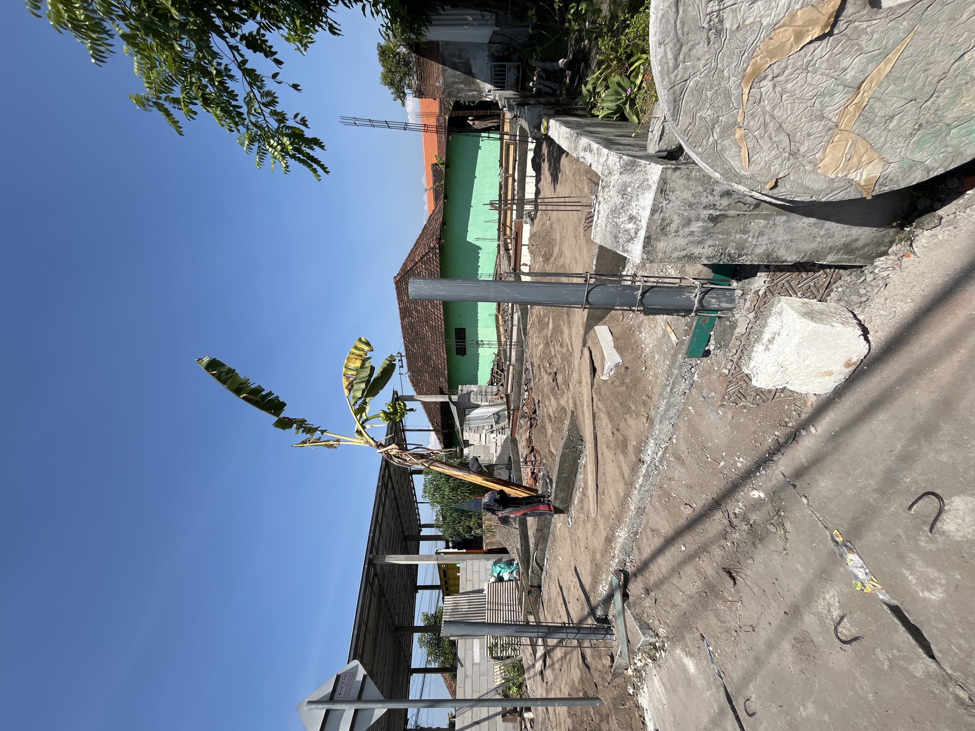 The image depicts a construction site in Pesawahan, Indonesia, on a clear, sunny day. The foreground shows a partially built structure with concrete foundations and rebar. A tall, gray pipe stands vertically, secured by metal straps. Adjacent to it is a rough concrete pillar and a large, circular concrete object with a textured surface and some paper-like debris attached. A pile of white building blocks sits on the ground.

In the mid-ground, the construction continues with exposed rebar and wooden forms for concrete pouring. A banana tree with yellowing leaves leans precariously in the center of the scene, with small green bananas visible on its stalk. A person wearing a dark shirt and red and black striped pants is visible near the banana tree, appearing to be working.

Further back, a long, covered structure with a metal roof supported by pillars is present, with stacks of building materials, including corrugated metal sheets and bags of what appears to be cement or sand, stored beneath it. Behind this, a long, green building with a red-tiled roof is visible, and to its right, a section of a brick wall and another building with a terracotta-colored roof. The environment is dusty and appears to be a typical urban or semi-urban construction area. The sky is a clear, vibrant blue.
