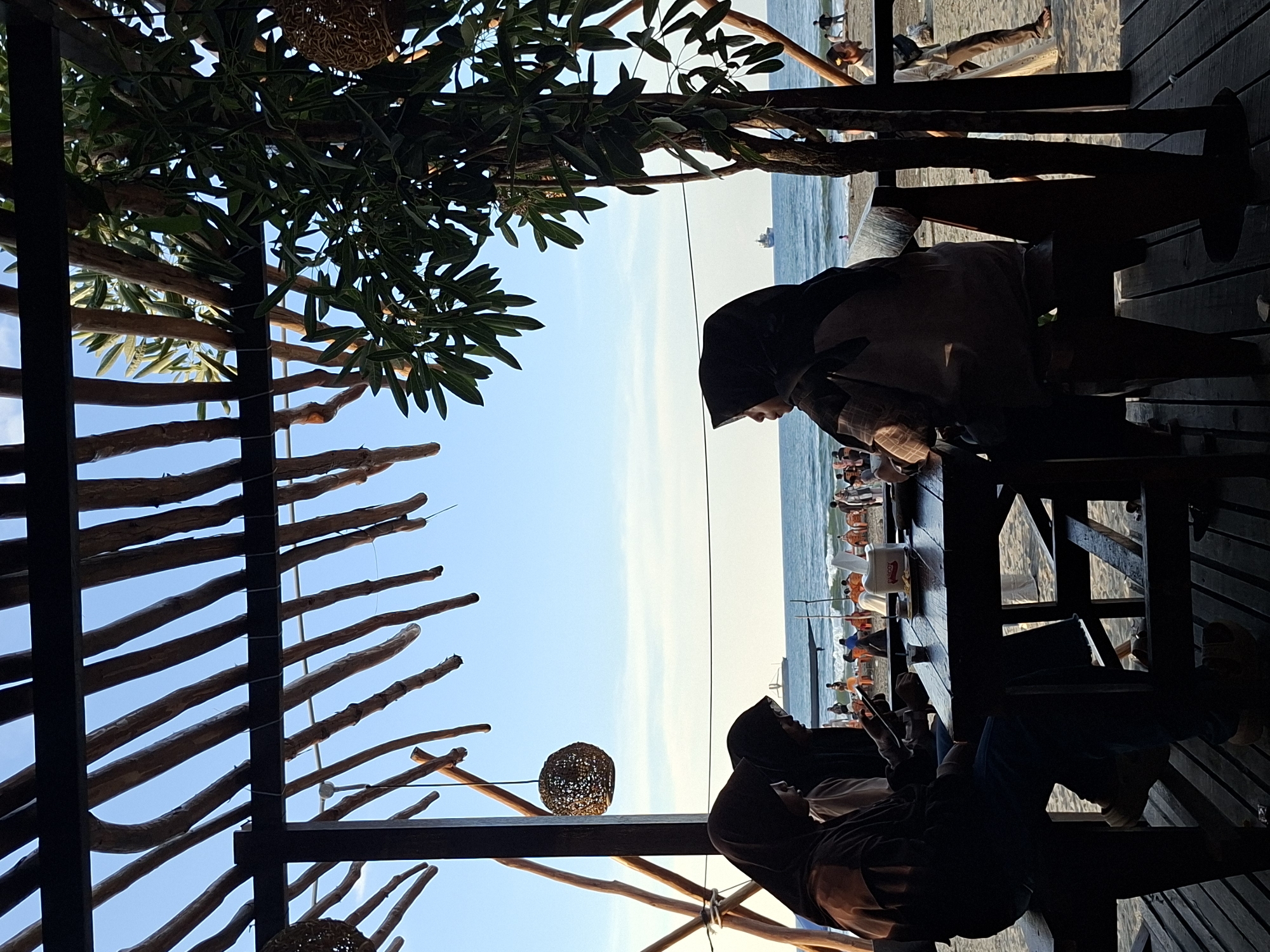 From a slightly low angle, the image depicts a beachside scene in Keudekruenggeukueh, Indonesia. Two individuals wearing hijabs are seated at a table, suggesting a cafe or restaurant setting. The environment includes elements of beachside infrastructure, such as a wooden structure overhead, possibly a pergola or awning, with branches and leaves providing partial shade. In the distance, the beach is visible, populated with people and objects associated with a coastal environment, like umbrellas, chairs, and the sea itself. A ship is seen near the horizon.