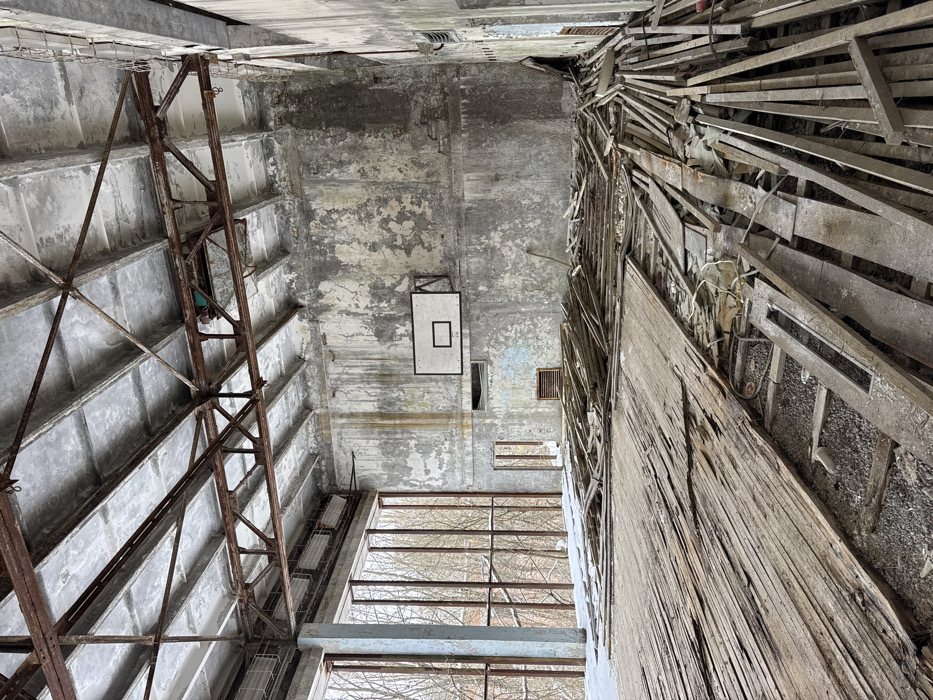 The image captures a top-down, slightly oblique view inside a severely dilapidated and abandoned building, likely a gymnasium or large hall, characterized by extensive decay and structural damage. The perspective looks downwards from a significant height, revealing multiple levels and the overall ruinous state of the interior.
On the left side of the frame, a large, rusty, metal skeletal structure descends diagonally. This appears to be a heavy-duty support beam or part of a large, industrial-style truss system, made of interconnected square and triangular metal bars, showing significant corrosion. Behind this structure, the ceiling or upper wall features a textured concrete surface with a grid pattern, displaying signs of peeling paint, water damage, and general grime.
The right side of the image is dominated by a massive, severely damaged wooden structure that also extends downwards diagonally. This appears to be the remnants of collapsed or decaying bleachers or tiered seating, with numerous wooden planks splintered, broken, and haphazardly piled. Some wires or cables are visible amidst the wooden debris, suggesting former electrical installations. The wall behind this wooden wreckage is equally deteriorated, with large sections of paint peeled away, exposing rough concrete or plaster.
In the central and lower portions of the image, towards what would be the far wall and floor of the building, the extent of the abandonment is clearly visible. High on a distant wall, a faded white rectangular panel with a black outline and a smaller black rectangle inside is prominently displayed, indicating a basketball backboard. The actual hoop is missing. Below this, further down the wall, several rectangular openings or vents are visible.
Towards the very bottom of the frame, large window openings reveal the exterior. Through these windows, a dense tangle of bare tree branches is visible, suggesting the image was taken during late autumn or winter when trees have shed their leaves. The overall color palette is dominated by muted grays, browns, and dull whites, reflecting the concrete, rust, and decayed wood, with minimal hints of faded color. The lighting appears to be natural, possibly from an overcast day, illuminating the scene from the window openings. The image conveys a strong sense of forgotten history, structural failure, and nature reclaiming what was once a functional space.