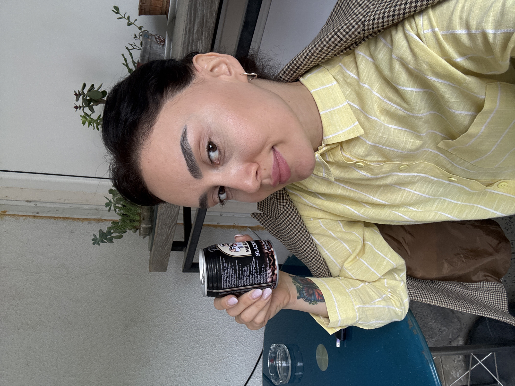 A woman with dark hair, wearing a yellow striped shirt and a brown checkered jacket, is holding a can of Mr. Brown Black Coffee. She has a butterfly tattoo on her arm. She is sitting at a blue table with an ashtray, seemingly outdoors in Tbilisi, Georgia, due to the Georgian text on the can. A plant is visible in the background.