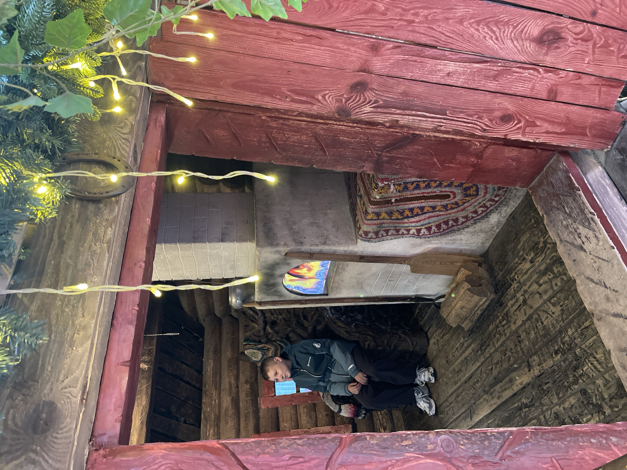 A slightly low-angle view of a wooden structure decorated with holiday lights, possibly an outdoor attraction in Moscow, Russia. Inside the structure, a boy wearing a grey and black jacket and black pants is lying on his side. His left cheek is partially obscured by a surgical mask. He is wearing white sneakers. The interior walls of the structure appear to be made of roughly hewn logs and there is a decorative rug hanging on an interior wall. A colorful, stained-glass window is visible in the background. The outer structure appears to be made of red-painted wooden planks. The view is partially obstructed by lit Christmas tree branches on the left side. A horseshoe is affixed to the wooden support.