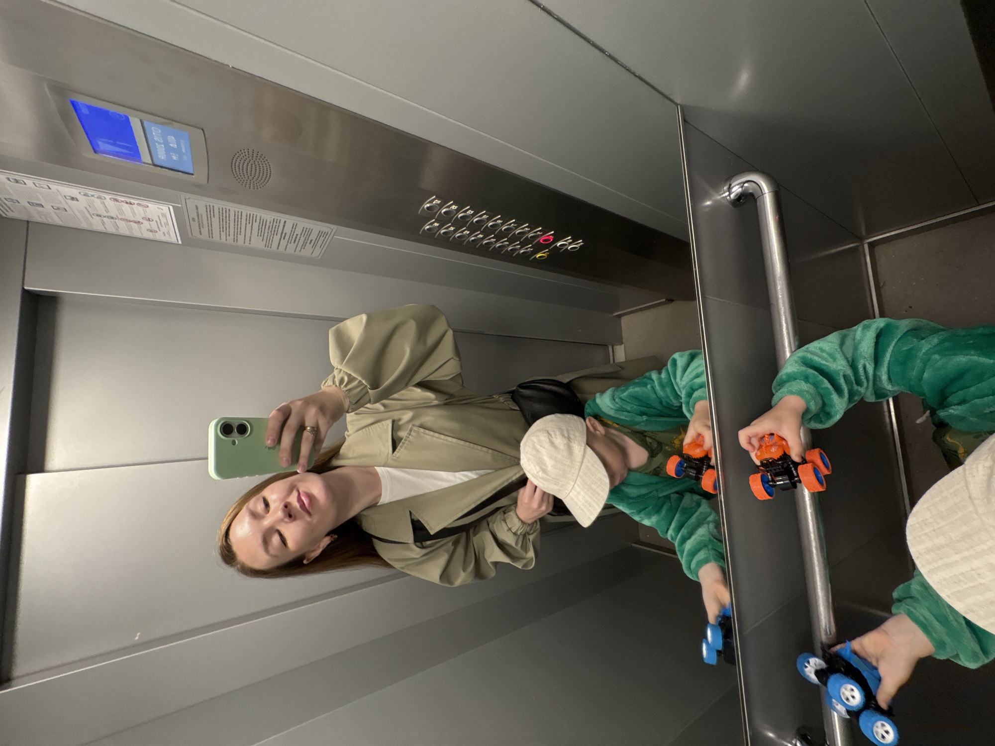 A woman is taking a selfie in an elevator with a child. The woman has long, light brown hair and is wearing a tan trench coat over a white t-shirt. She is holding a mint green smartphone in her right hand and is wearing a ring on her left ring finger. The child is wearing a beige baseball cap and a green hooded jumpsuit. The child is holding two toy cars, one blue and one orange. The elevator has stainless steel walls and a silver handrail. The elevator panel has buttons for floors 1 through 12, with floors 1 and 2 illuminated in red. There is also a digital display showing "ANOS ZITO" and other text. The image appears to be taken in a residential building in Izhevsk, Russia.