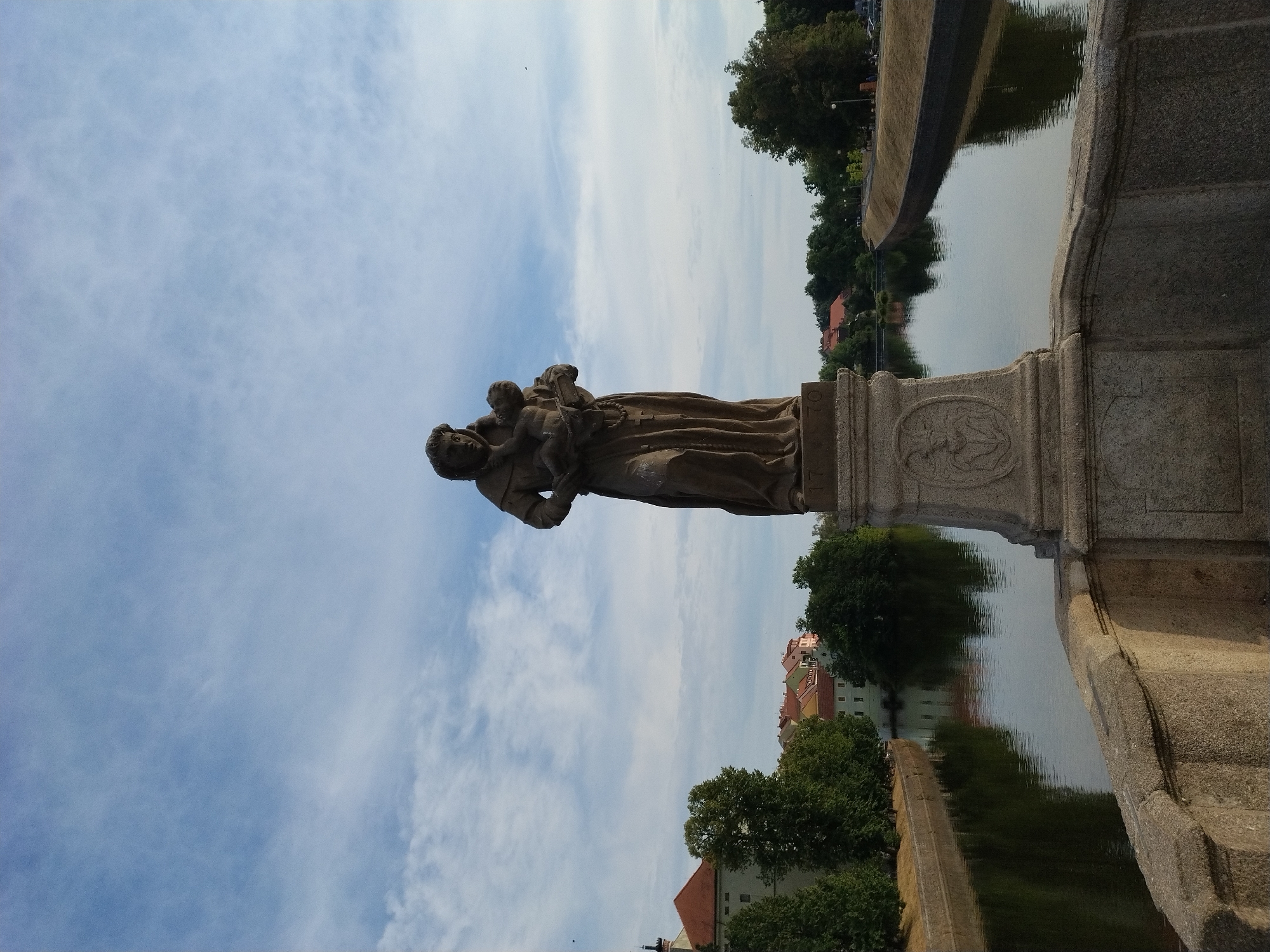 This image depicts the Saint John of Nepomuk statue, a notable landmark situated on the Písek Bridge in Písek, Czechia. The stone statue, carved in Baroque style, portrays Saint John of Nepomuk holding the infant Jesus in his left arm and a crucifix in his right. The statue stands on a pedestal adorned with bas-reliefs. The bridge itself is constructed from stone, with a balustrade visible in the foreground. The statue overlooks the Otava River, with reflections of the surrounding architecture and lush green trees visible on its calm surface. The cityscape in the background features traditional Central European buildings with red-tiled roofs, characteristic of Písek. The sky above is a pale blue with scattered white clouds, indicating fair weather.