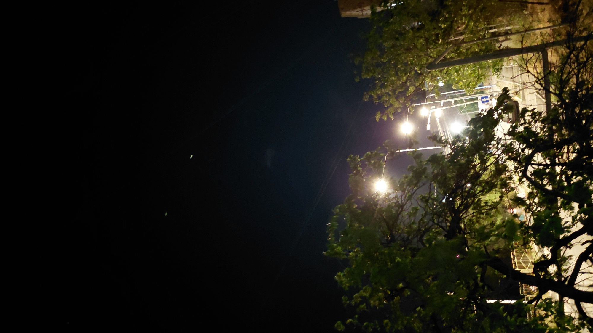 The image is a nighttime photograph taken in Abovyan, Armenia. The foreground is dominated by the dark silhouettes of trees with green leaves, illuminated by streetlights. In the background, several streetlights cast a bright glow, illuminating a road and what appears to be a building. There are faint streaks of light in the sky, possibly from distant lights or atmospheric phenomena. There are no visible people or animals in the image.
