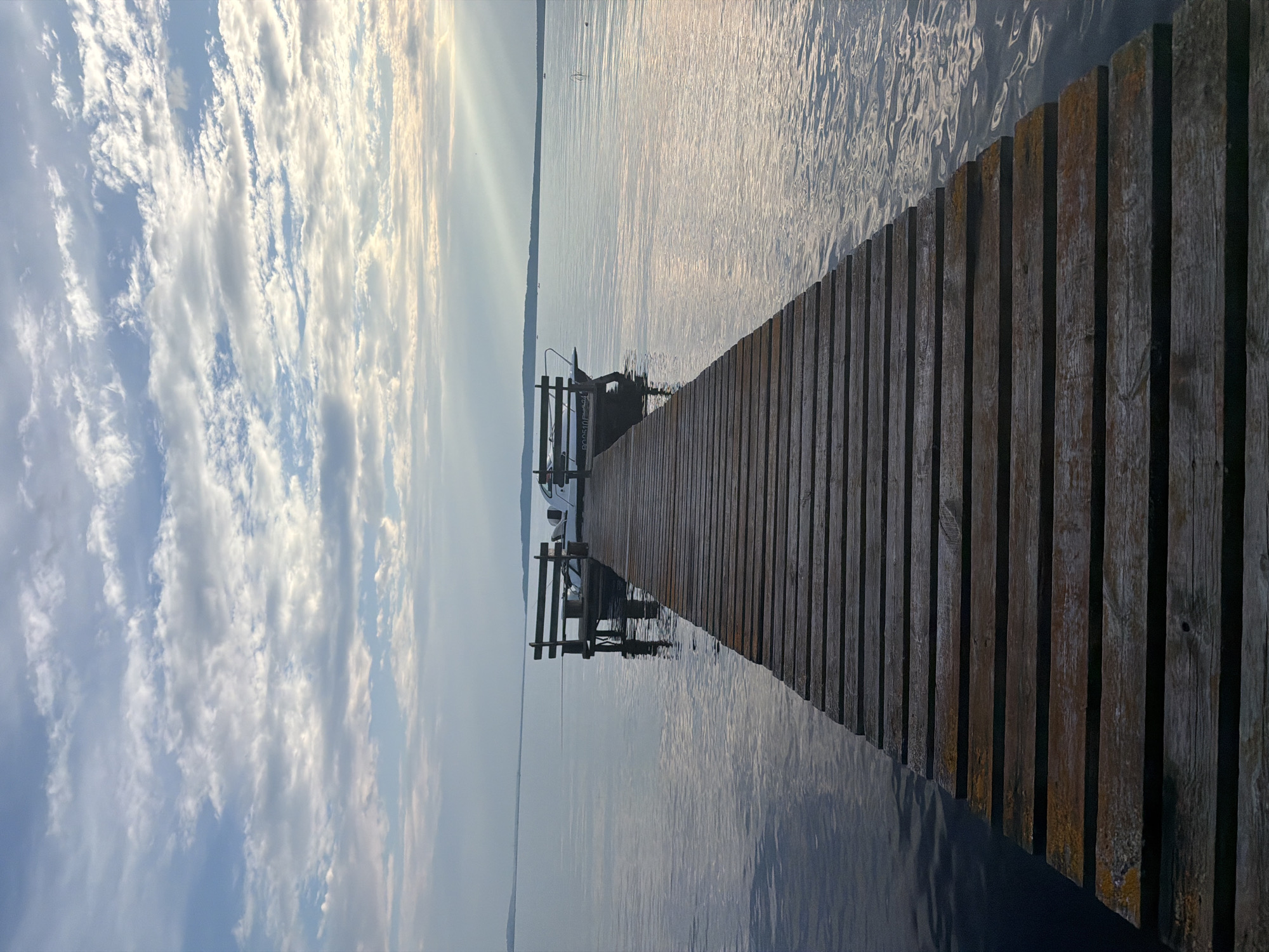 The image depicts a wooden pier extending into a calm body of water, likely a lake or bay. The pier is made of weathered wooden planks laid horizontally, creating a linear perspective that leads the eye towards the end. At the end of the pier, there are two wooden benches and a small white boat moored. The water is still, reflecting the soft light of the sky. The sky itself is filled with scattered clouds, some of which are illuminated by sunlight breaking through. The overall atmosphere is serene and peaceful. The location is identified as Stepanovo, Russia, suggesting a possibly rural or recreational setting. There are no people visible in the image.
