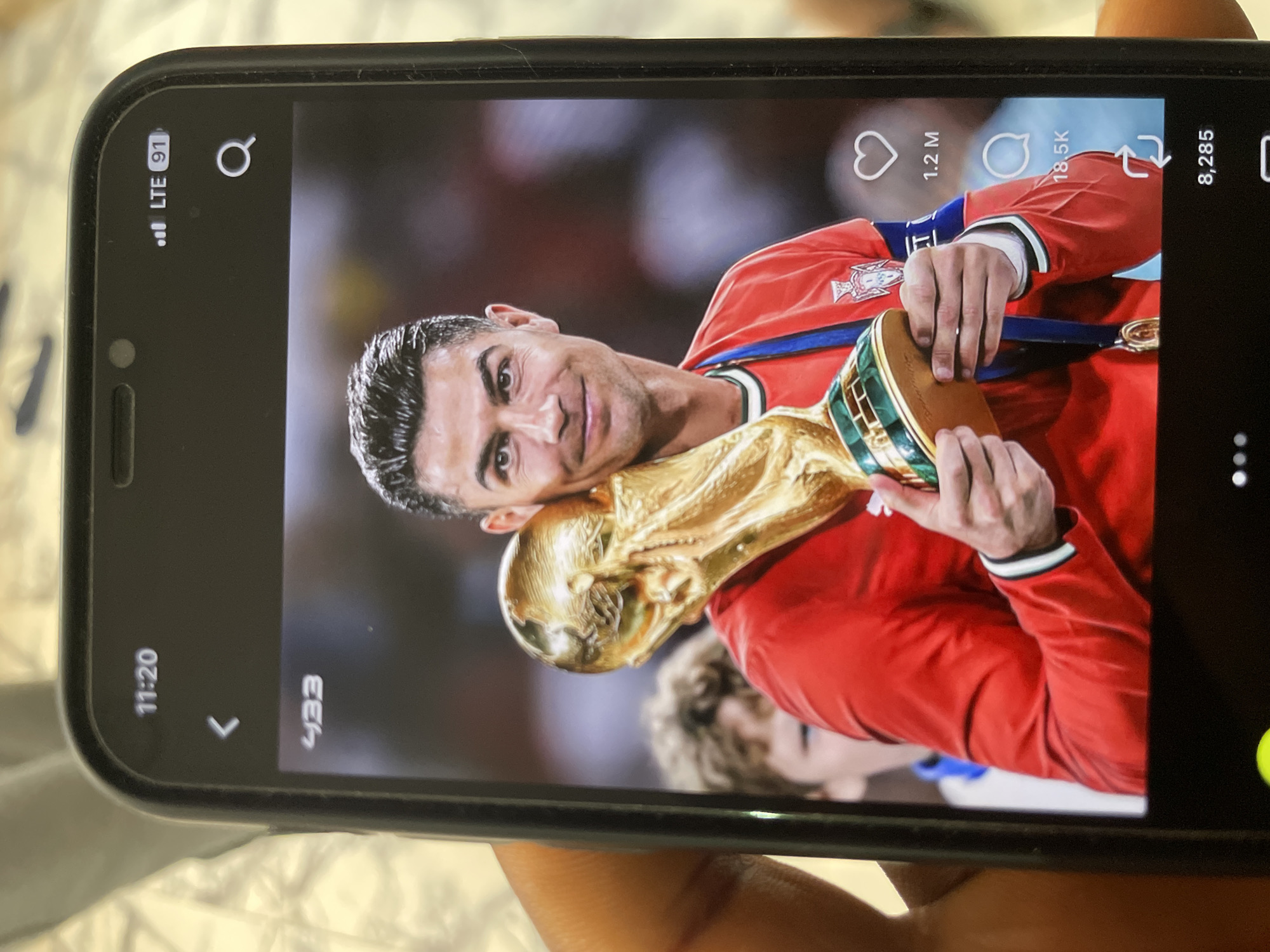A phone displays an image of Cristiano Ronaldo holding a trophy. He is wearing a red soccer jersey with a blue ribbon around his neck. The trophy is golden and appears to be the FIFA World Cup. Ronaldo is smiling at the camera. The image is displayed on a smartphone, held by a person whose hand is partially visible. The phone's interface shows a timestamp of 11:20, a back arrow, the account name "433", LTE signal strength, and icons for likes, comments, and shares. The background of the image on the phone is blurred, showing what appears to be a crowd. The phone itself is black and has a prominent notch at the top.