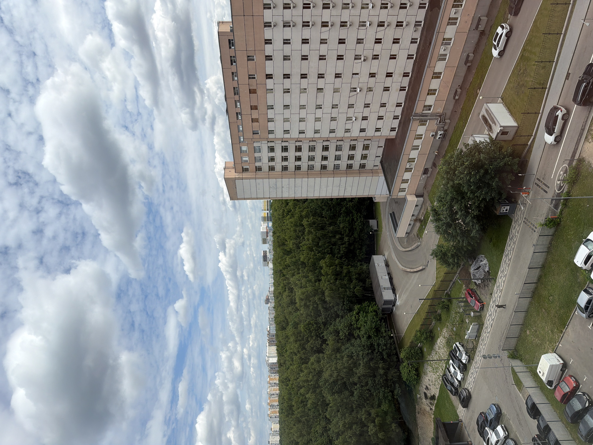 This is a wide-angle, high-angle aerial photograph of a residential area in Moscow, Russia. In the foreground, a tall, multi-story apartment building dominates the right side of the frame. It is light-colored with beige trim and numerous windows, along with several air conditioning units visible on its facade. A smaller building complex runs along the lower portion of this building.
To the left of the apartment building, a dense, green forest stretches into the background. Beyond the forest, a cityscape of numerous apartment buildings can be seen under a cloudy sky with patches of blue. The sky is filled with dramatic, white cumulus clouds.
In the mid-ground, a paved road curves around the apartment building, with several parked cars visible. One of these cars is red, and there is also a white utility van parked nearby. A modular structure, possibly a small office or storage unit, sits alongside the road.
Further left, in the foreground, a parking lot is filled with various vehicles, including sedans, SUVs, and what appears to be a white van. A pedestrian is walking on a paved path in front of a chain-link fence, near a red car. The ground is mostly covered in grass and some paved areas. The overall impression is an urban environment with significant green space.