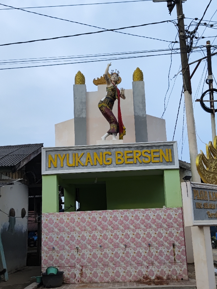 The image captures an outdoor scene in Kecamatan Selagai Lingga, Indonesia, likely during the daytime under an overcast sky. The central subject is a statue of a female dancer in traditional attire, adorned with a golden crown, armbands, and a flowing red sash, balanced on one leg with arms raised. The statue is positioned atop a light-colored structure featuring two golden, spire-like ornaments.
Below the statue, a building with a light green facade bears a prominent sign with the yellow text "NYUKANG BERSENI". Below this sign, a wall is covered in a decorative pattern of pink and white floral tiles, with several water spigots protruding from it. On the ground in front of this wall, a black bucket and a green bowl are visible.
To the right of the central structure, a utility pole with a dense network of black electrical wires extends upwards, crossing the upper portion of the frame. Part of another sign is visible on the far right, partially obscured, with the text "KEC. SELAGAI LINGGA" decipherable. On the left side, a section of a building with a dark, corrugated roof and a light-colored wall can be seen. The overall impression is of a vibrant local landmark or community facility.