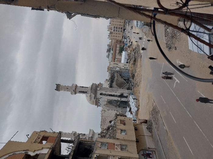The image depicts a heavily damaged urban street scene in Khan Yunis, Palestinian Territory, under an overcast sky. In the center, a large mosque with a prominent, partially intact minaret stands severely damaged, its dome and upper structure largely collapsed into rubble. To the immediate left, a multi-story building exhibits extensive structural damage, with walls blown out, exposed interiors, and debris scattered below. Another damaged building facade is visible on the far right, partially obscured by a dark metal railing in the foreground, suggesting an elevated vantage point for the photograph.
The paved road in the foreground and mid-ground shows visible white lane markings and arrow symbols. Numerous individuals, approximately 15-20, are observed walking along the road and its debris-strewn verges. Three individuals are walking together in the mid-left, while a person in a red cap stands in the lower-left foreground. Several others are distributed further down the street. A white van is parked on the right side of the road in the mid-distance. The surrounding environment is characterized by widespread destruction, with significant piles of rubble, exposed building skeletons, and scattered debris visible throughout the scene. What appear to be makeshift shelters or structures are discernible near the damaged mosque.
