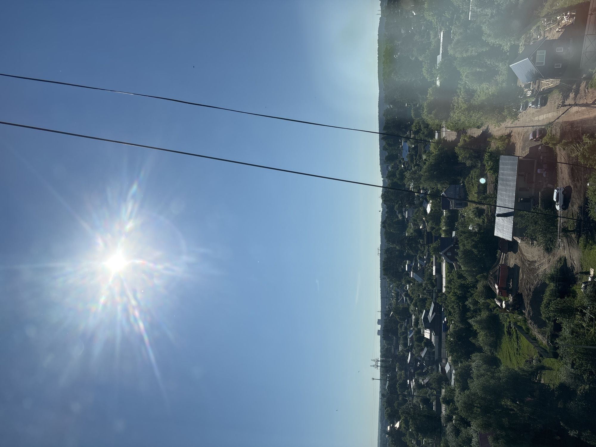 This is an aerial view of a suburban area in Chaikovsky, Russia, on a sunny day. The sun is bright in the upper left quadrant of the image, creating lens flare and a starburst effect. Two thick, dark cables run diagonally from the top right to the bottom left, indicating the perspective is from a cable car or similar elevated structure.
Below the sky, a dense canopy of green trees covers the landscape, interspersed with residential buildings. The houses appear to be of varying sizes and styles, many with dark roofs, and some are surrounded by fences. Several dirt roads wind through the community.
In the midground, on a patch of disturbed earth, are construction vehicles: a yellow backhoe, an orange dump truck, and a smaller truck. Piles of dirt and construction materials are visible around them. Further in the distance, power lines and transmission towers are visible against the horizon, along with some taller, box-like structures. The overall impression is of a developing or established residential area with ongoing construction activity, under a clear blue sky. There are no visible people in the frame.