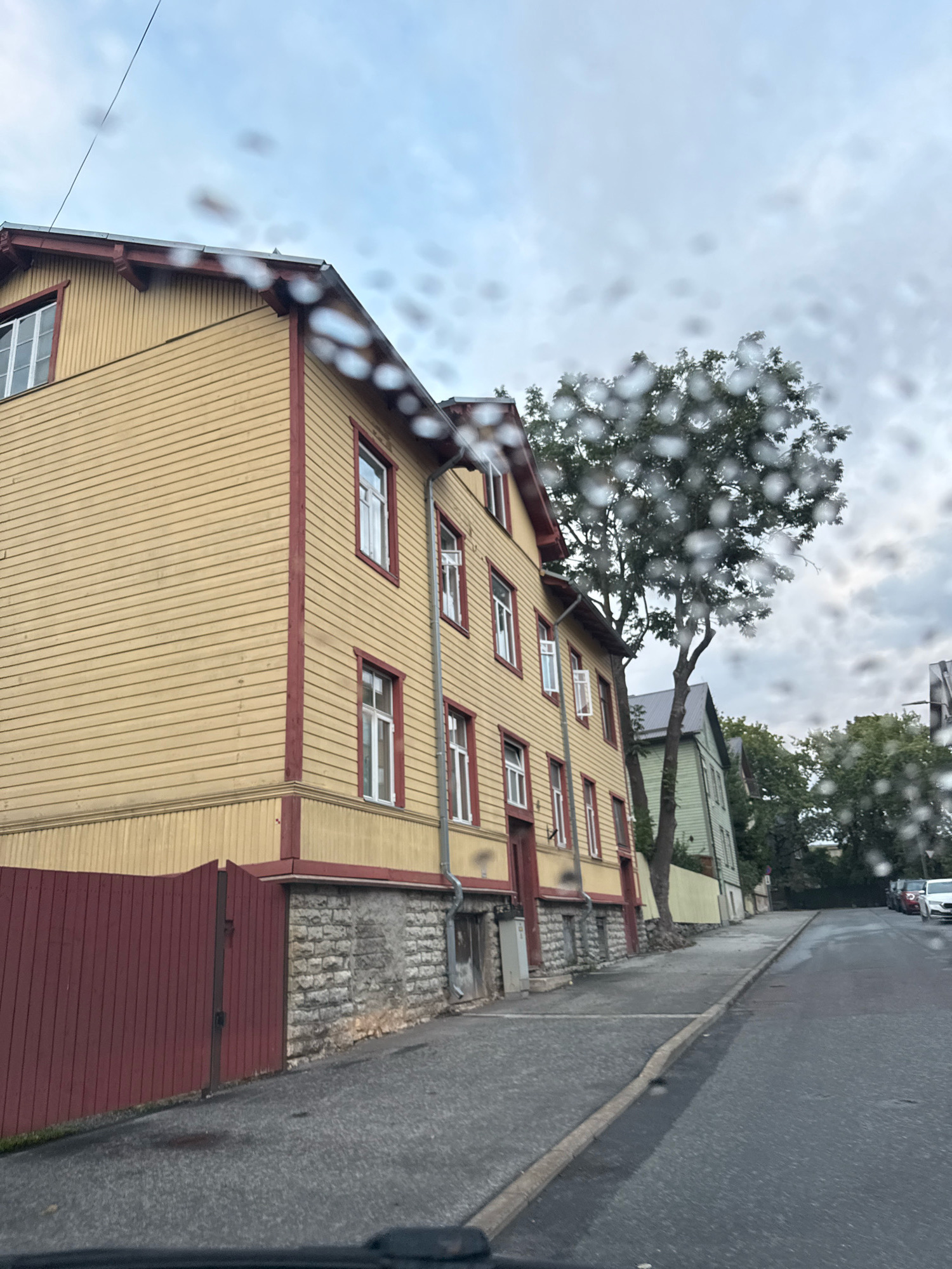 The image shows a street scene in Tallinn, Estonia, on a cloudy day with raindrops on the lens. A prominent yellow wooden building with red trim and numerous white-paned windows lines the left side of the street. In front of the building, there is a dark red wooden fence. The street is paved and appears wet, with a sidewalk to the left of the road. Several trees are visible, including a large deciduous tree partially obscuring the view of another green building further down the street. A few parked cars are visible in the distance on the right side of the road. The overall impression is of a residential street in a European city, with the raindrops adding a sense of atmosphere.