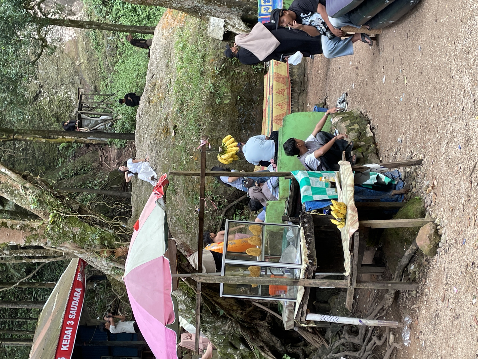 The image depicts an outdoor scene likely near a natural attraction, possibly a waterfall or cave, given the lush greenery and rocky terrain. In the foreground, a small food stall with a pink and white umbrella displays fried food items in a glass case. A man is sitting to the left of the stall, and another man with a cigarette is seated to the right, wearing a black t-shirt and jeans.
In the middle ground, several people are gathered. One individual in a white dress is seated on a large rock, posing for a photograph. Others are standing or sitting, with some appearing to be vendors or patrons. A bunch of bananas hangs from a stall.
In the background, a weathered wooden structure with a blue and red sign that reads "KEDAI 3 SAUDARA" (which translates to "3 Siblings Shop") is visible. More people are seen further up a path leading to a higher elevation. The overall environment is characterized by dense vegetation, large trees, and rocks, suggesting a rural or semi-rural setting. The presence of a shop sign indicates a locality, potentially within or near a tourist area. While the image itself does not explicitly state the location as Sarilamak, Indonesia, it captures a scene consistent with such a region, known for its natural beauty and local businesses catering to visitors.