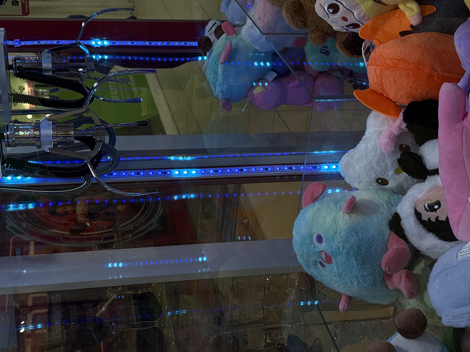 The image shows a claw crane game filled with plush toys. The game is illuminated by blue LED lights. The crane's mechanism is visible, consisting of metal arms and a claw. A variety of plush toys are crammed inside, including a blue plush character with pink features, an orange plush toy resembling a pumpkin, a brown plush figure, and other assorted stuffed animals. The glass enclosure of the game reflects the surrounding environment. There is no explicit indication of location, people, or activity within the game itself.