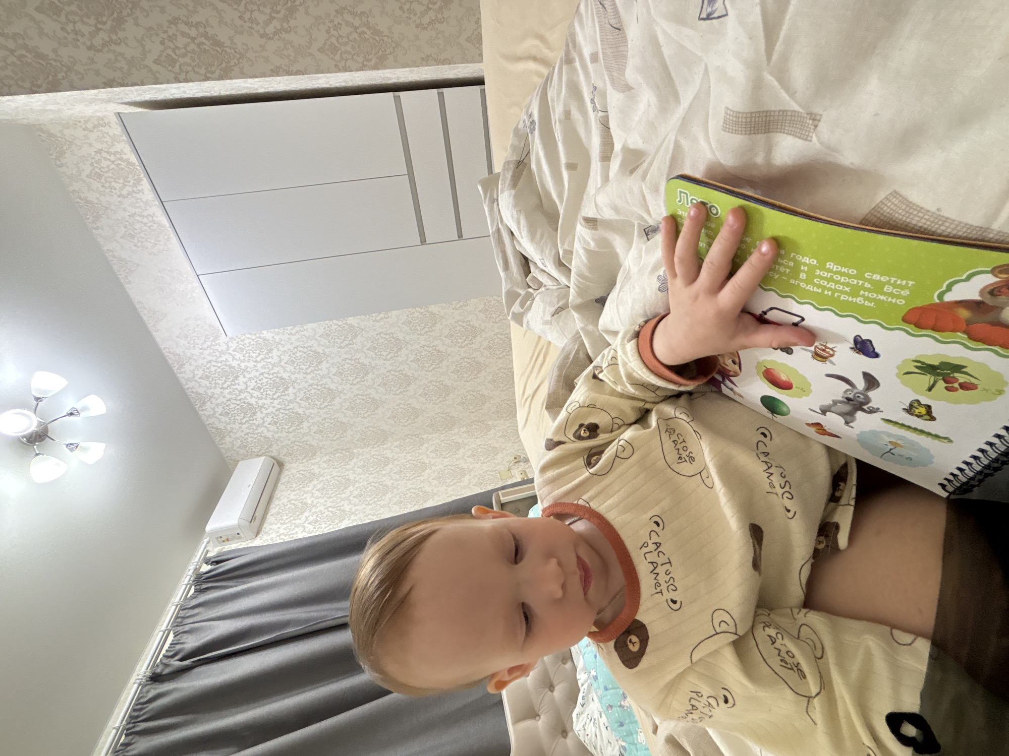 Here is a description of the image:
A baby wearing a brown and white printed pajama is looking at a book with a green cover. The book has colorful images on it. The baby's hand is resting on the pages of the book. The background shows a bedroom setting with a bed covered in patterned sheets, a white cabinet, a curtain, and a ceiling light. The location is likely in a home, possibly in Ферма, Russia, as indicated by the language on the book.