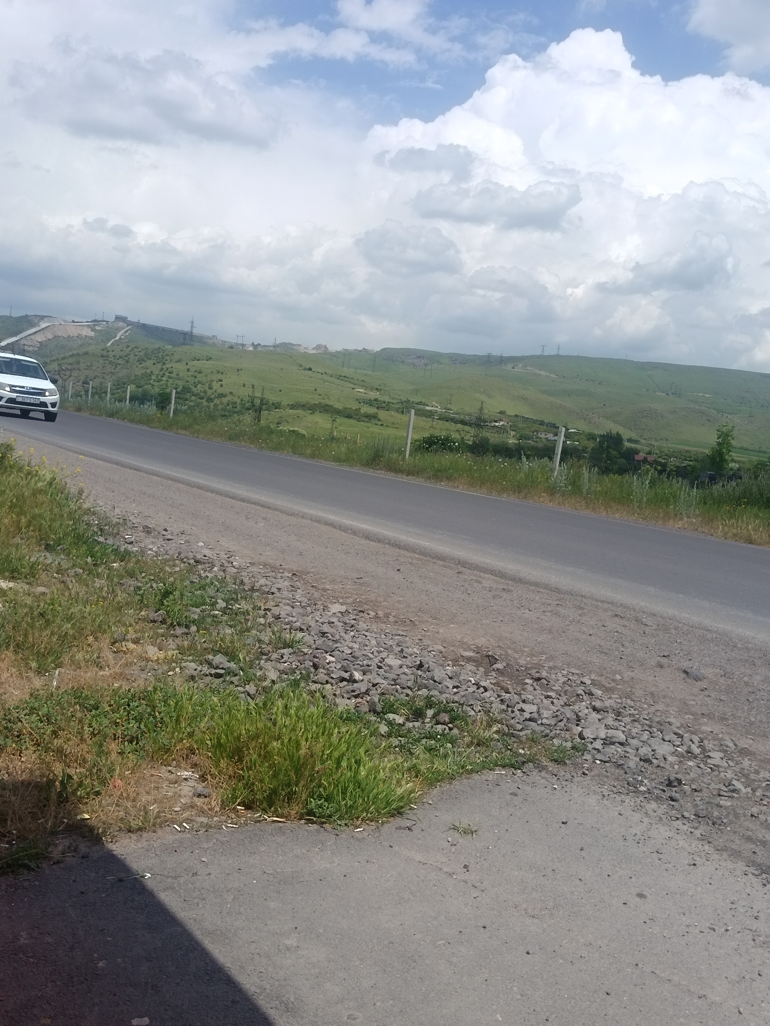 An eye-level, wide shot captures a paved road curving through a hilly, green landscape under a partly cloudy sky. A white car drives away from the viewer on the road, its license plate visible. The road is bordered by a gravel shoulder with patches of dry and green grass. In the background, rolling hills covered in green vegetation are dotted with trees and buildings, with power lines visible on the distant slopes. The sky is a mix of bright blue and large, fluffy white clouds.