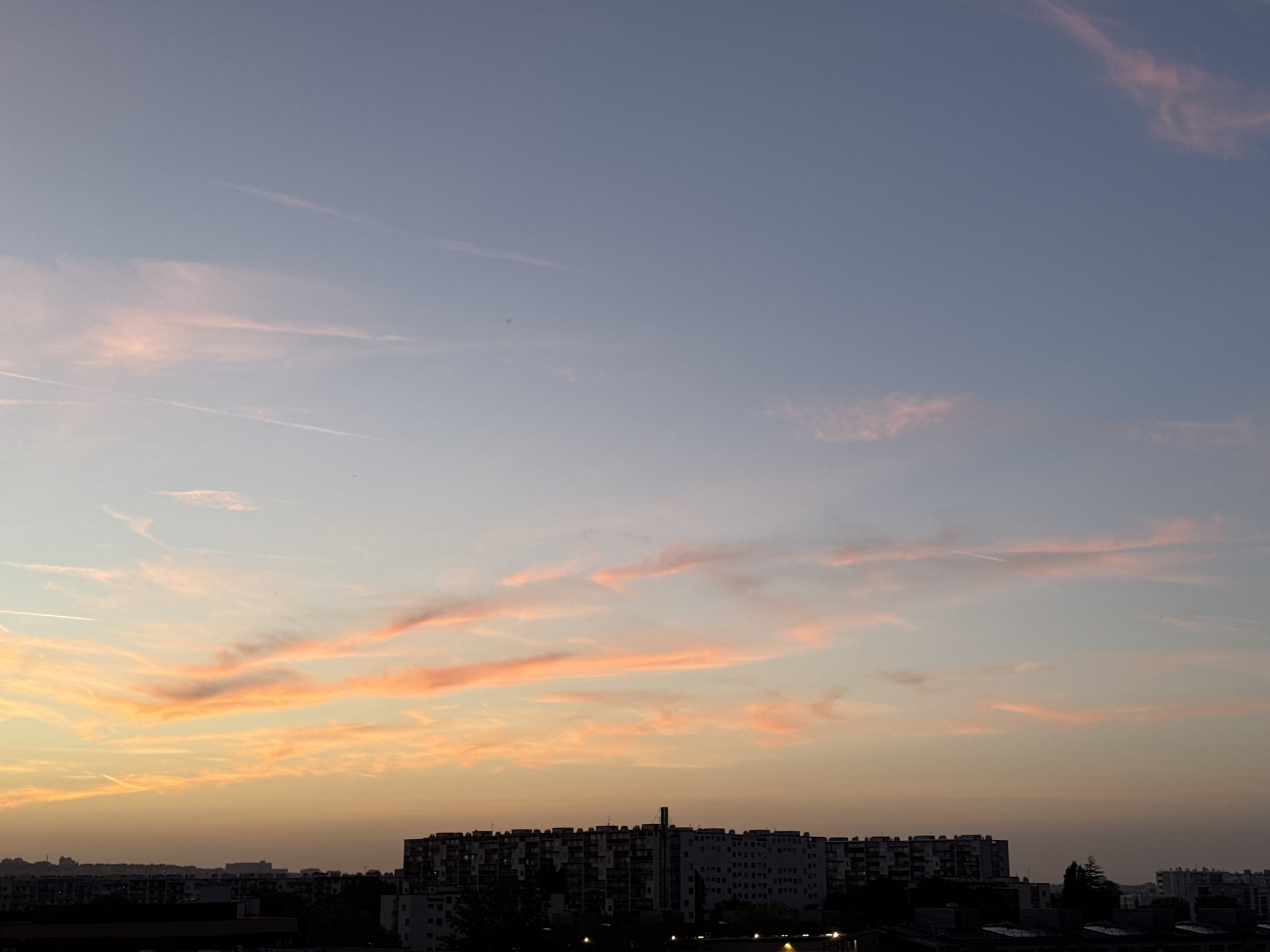 The image depicts a city skyline at dusk. The sky is a gradient of soft blues and oranges, with wispy pink clouds scattered throughout. Contrails from airplanes are visible in the upper portion of the sky. In the foreground, a silhouetted cityscape stretches across the horizon, characterized by rows of apartment buildings and other structures. The lighting suggests the sun has recently set, casting a warm glow on the clouds and illuminating the scene. The overall impression is one of a tranquil evening in an urban environment.