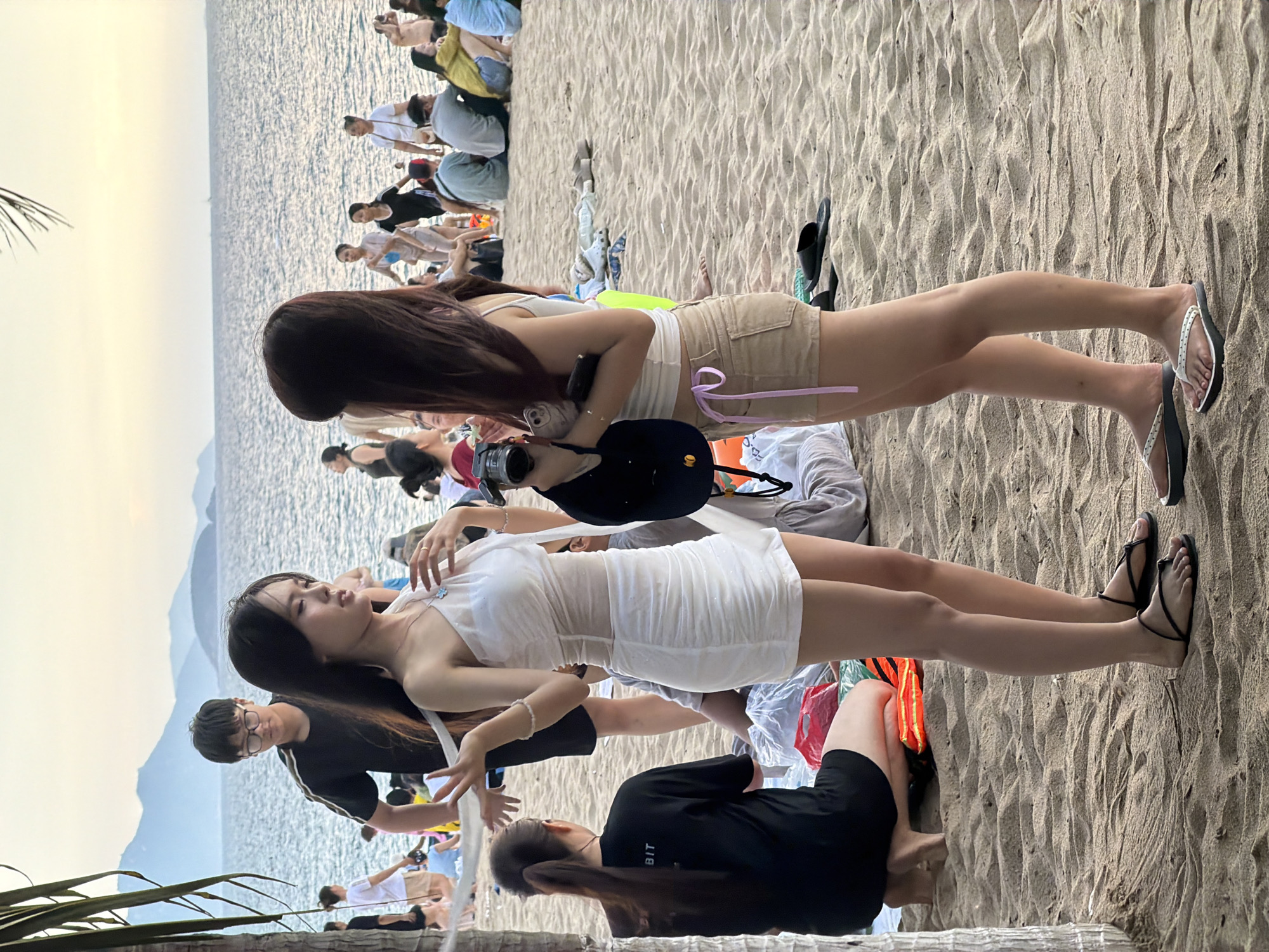 This media file depicts a group of people on a sandy beach, with the ocean and mountains in the background. The image is oriented vertically.
The main subjects are several individuals, primarily young women, dressed in casual beach attire. One woman in the foreground is wearing a white, form-fitting dress, and another is in khaki shorts and a white top. There are also people in the background, some of whom appear to be sitting or standing, suggesting a gathering or an event.
The scene appears to be a public beach, possibly a tourist area in Nha Trang Ward, Vietnam, given the context. The time of day seems to be late afternoon or early evening, as indicated by the soft lighting and the warm tones in the sky. The weather appears to be clear and calm.
Notable details include the sandy texture of the beach, the rippling surface of the water, and the distant outline of mountains. Some individuals are holding phones or cameras, suggesting they are taking pictures or documenting their experience. There is a sense of leisure and social interaction among the people.
One visible detail on clothing is the word "BIT" on the back of a black t-shirt worn by someone in the foreground. Overall, the media file captures a relaxed moment on a beach with a group of people enjoying the scenery and each other's company.