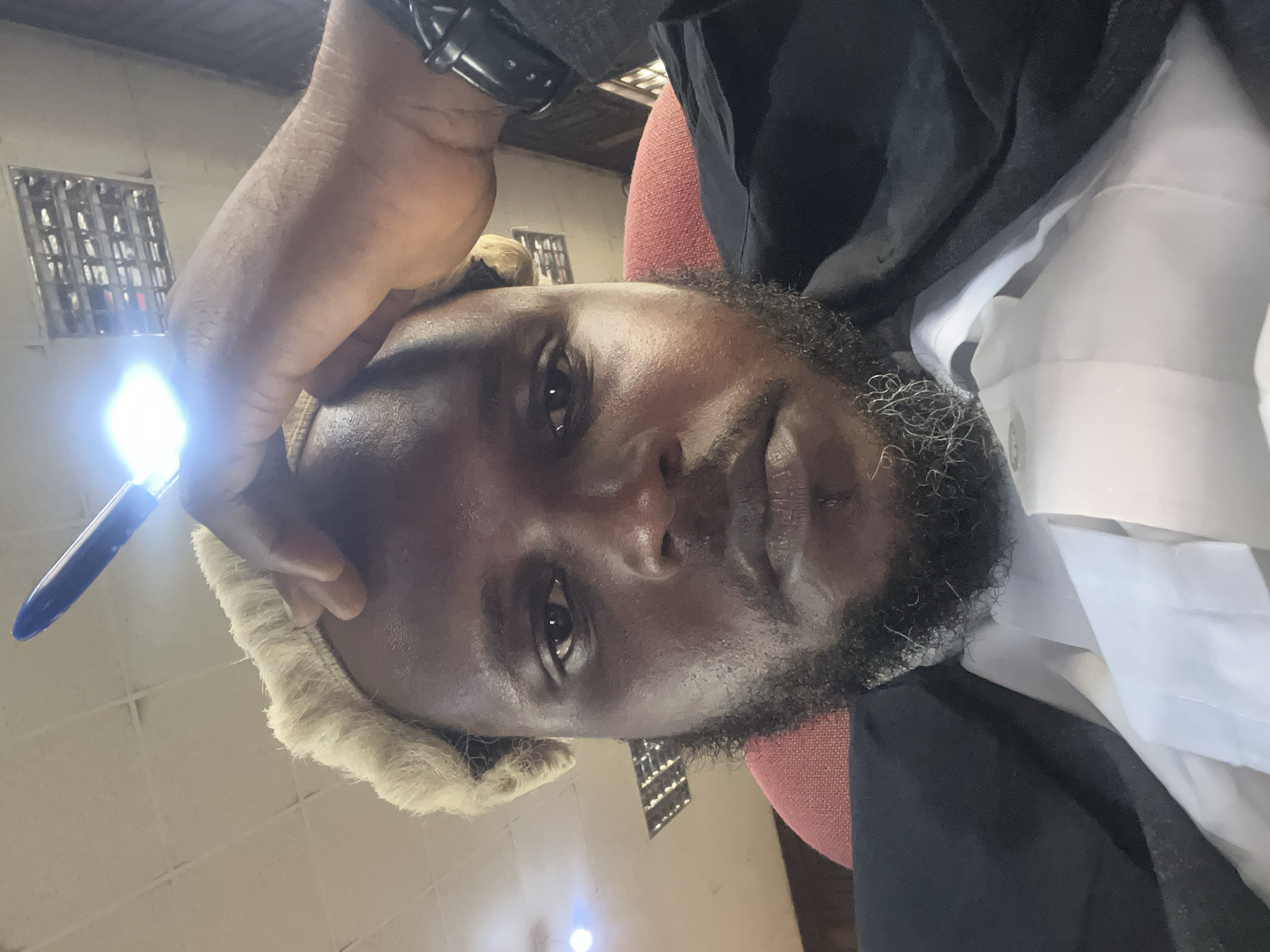 A close-up, slightly upward-angled selfie features a man with dark skin and a beard, positioned indoors in what appears to be a legal setting in Abeere, Nigeria. He wears a white collared shirt and a dark jacket or robe, along with a light-colored, possibly white or cream, legal wig. His expression is neutral and contemplative.
His left hand is raised, holding an object that emits a bright, intense light, possibly a pen with a built-in flashlight, positioned near his temple. A dark watch is visible on his left wrist. Behind him, a partial view of a red or maroon chair back is seen. The background includes a light-colored ceiling with visible paneling or a grid structure. To the left, a window with metal security bars or a distinct grid pattern is visible. Another diffuse light source appears in the lower right of the frame. The attire and setting strongly suggest he is a legal professional in a courtroom or office. No text is visible in the image.