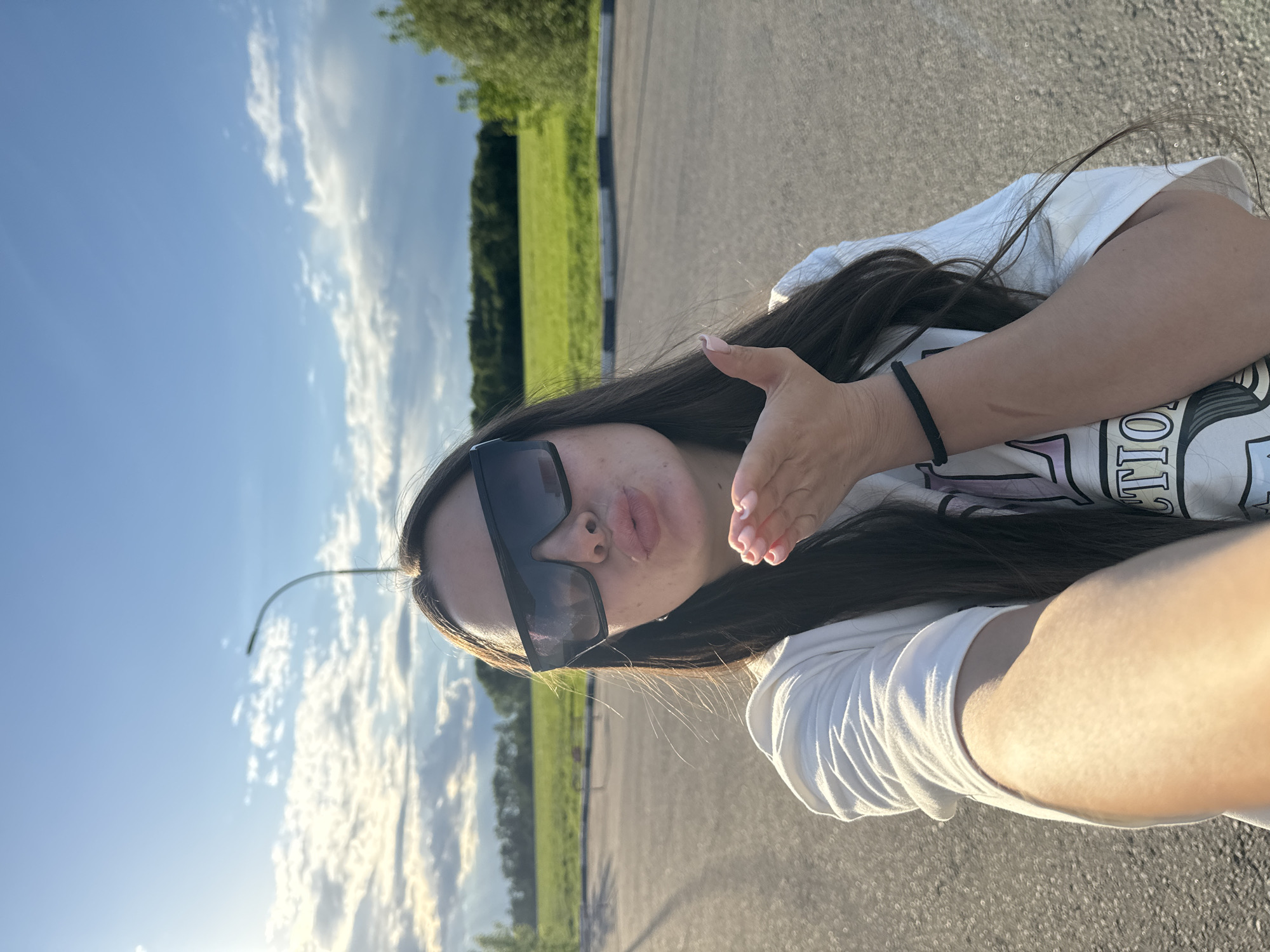 A young woman with long, dark hair and sunglasses is taking a selfie on an asphalt road. She is wearing a white t-shirt. She is outdoors, with a green field and trees in the background. The sky is blue with scattered clouds. The scene is in Bolshiye Vyazyomy, Russia. The woman is making a kissing gesture with her lips and hand. A street light is visible in the background.