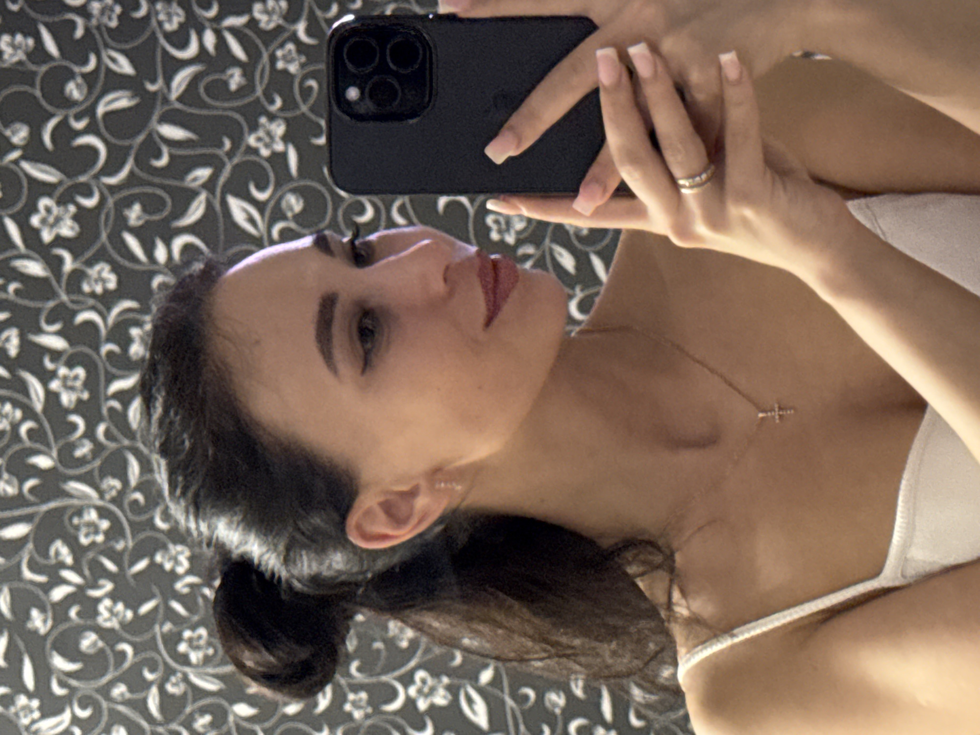 A woman with dark hair pulled back in a bun is holding a black smartphone to take a selfie. She is wearing a white top with thin straps and a gold necklace with a cross pendant. A wedding ring is visible on her finger. The background is a gray wall with a white floral pattern. The photo was most likely taken indoors. There is no information that indicates this photo was taken in Chaikovsky, Russia.