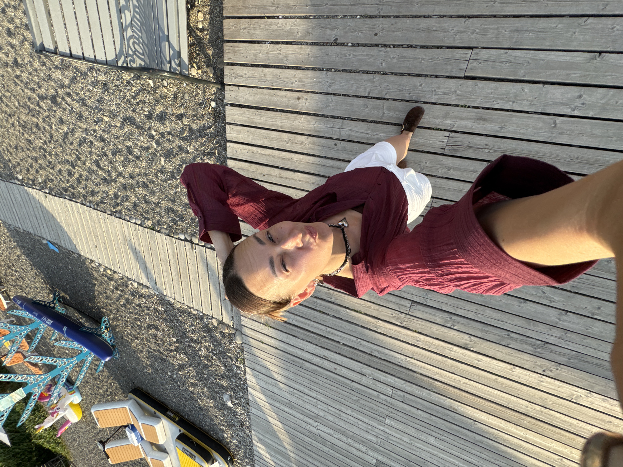 A woman is taking a selfie outdoors. She is standing on a wooden boardwalk, with a sandy area to her left and a light-colored bench in the distance to her right. The woman is wearing a maroon, long-sleeved, open-collared shirt and white shorts. She has a dark necklace with a heart-shaped pendant. Her right hand is resting on the back of her head, and she is winking at the camera with her eyes. Behind her, to the left, are some decorative elements that appear to be made of blue and white coral-like structures with various colorful objects attached, including what looks like a blue inflatable whale. Further left, partially visible, is a yellow and white inflatable boat. The overall setting suggests a sunny day near a body of water, possibly a beach or a pier area. The scene is likely in Sochi, Russia, given the presence of beach-related items and the time of year inferred from the attire and sunlight.
