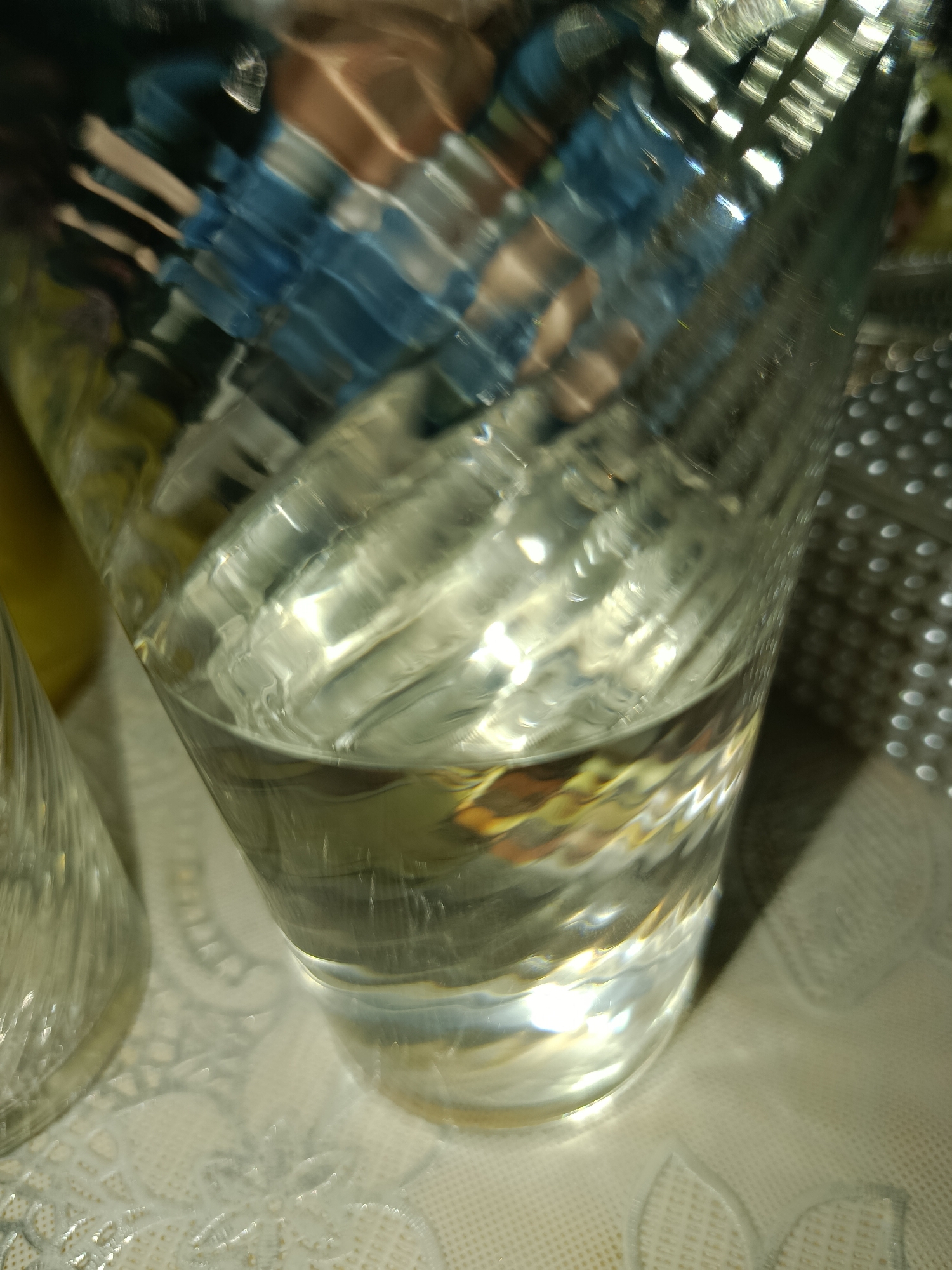 The image shows a close-up of a glass with water on a tablecloth. The glass has a textured surface that distorts the reflection of objects behind it, including what appears to be people wearing blue clothing. The tablecloth has a lace-like pattern. The scene is likely indoors, given the lighting and the presence of a tablecloth. The location is not specified in the image or context.